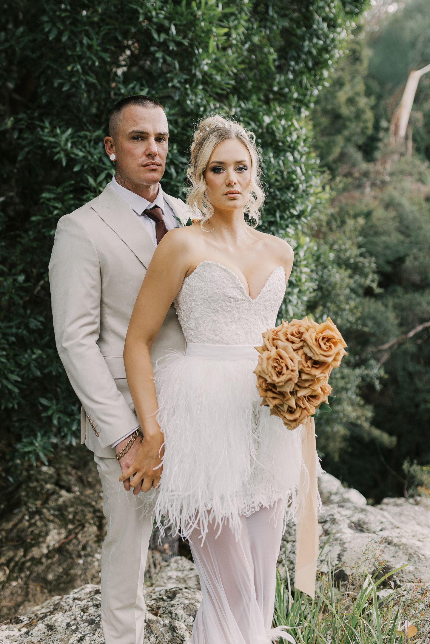 Bride in feathered white gown holding orange roses bouquet stands with groom in beige suit outdoors.