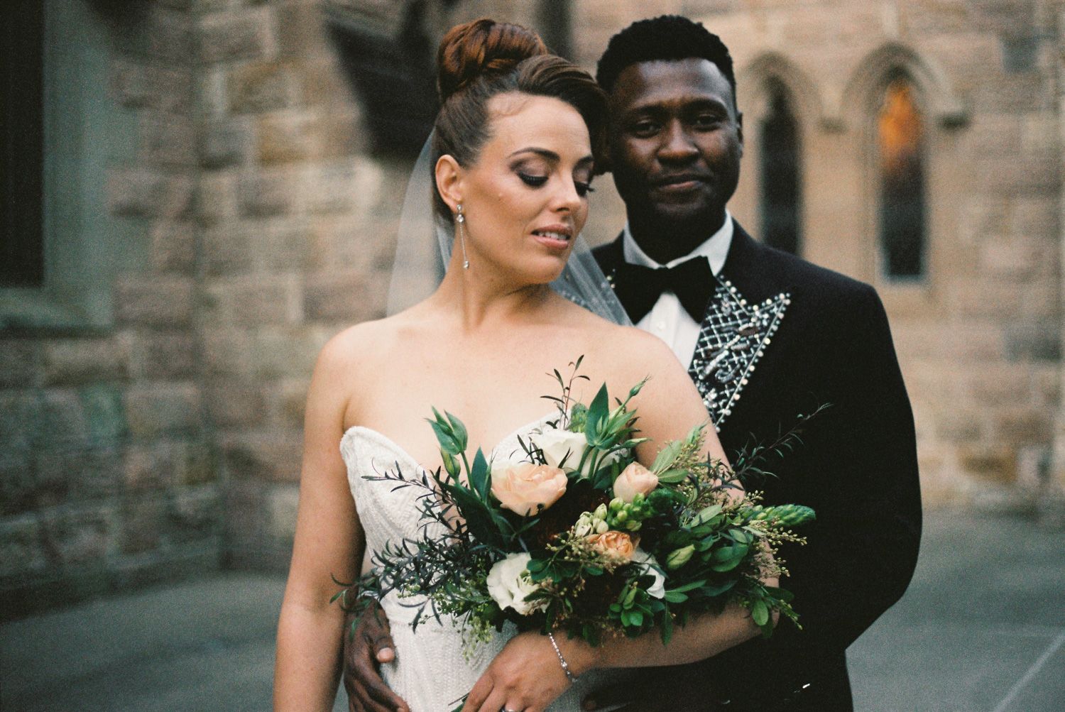 Bride and groom posing outdoors, bride holding a lush bouquet of roses and greenery, groom in a black tuxedo.