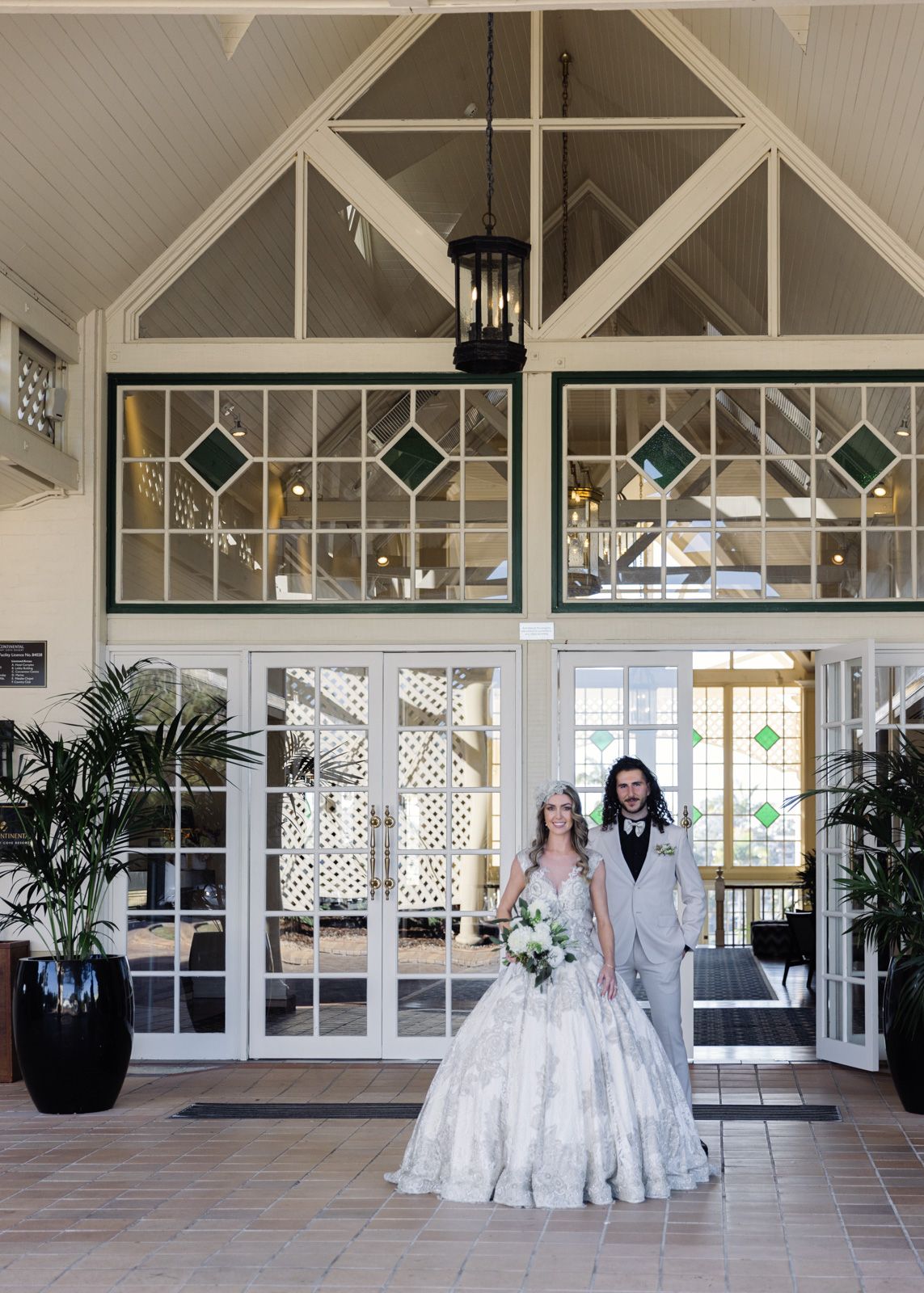 A wedding couple poses in front of a grand entrance with glass panels and architectural details.