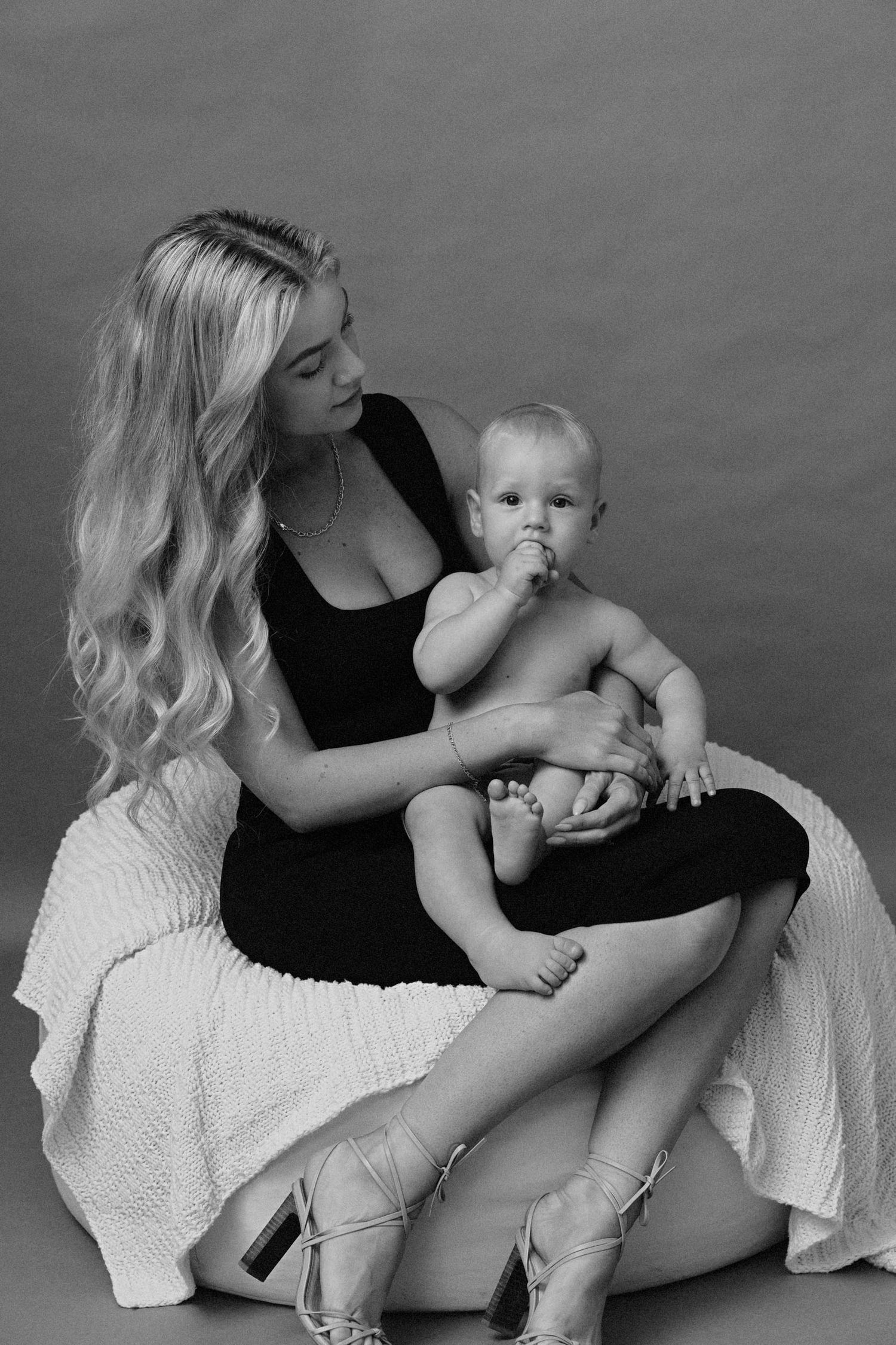 A black and white portrait of a parent and baby sitting together on a draped surface, wearing ballet shoes.