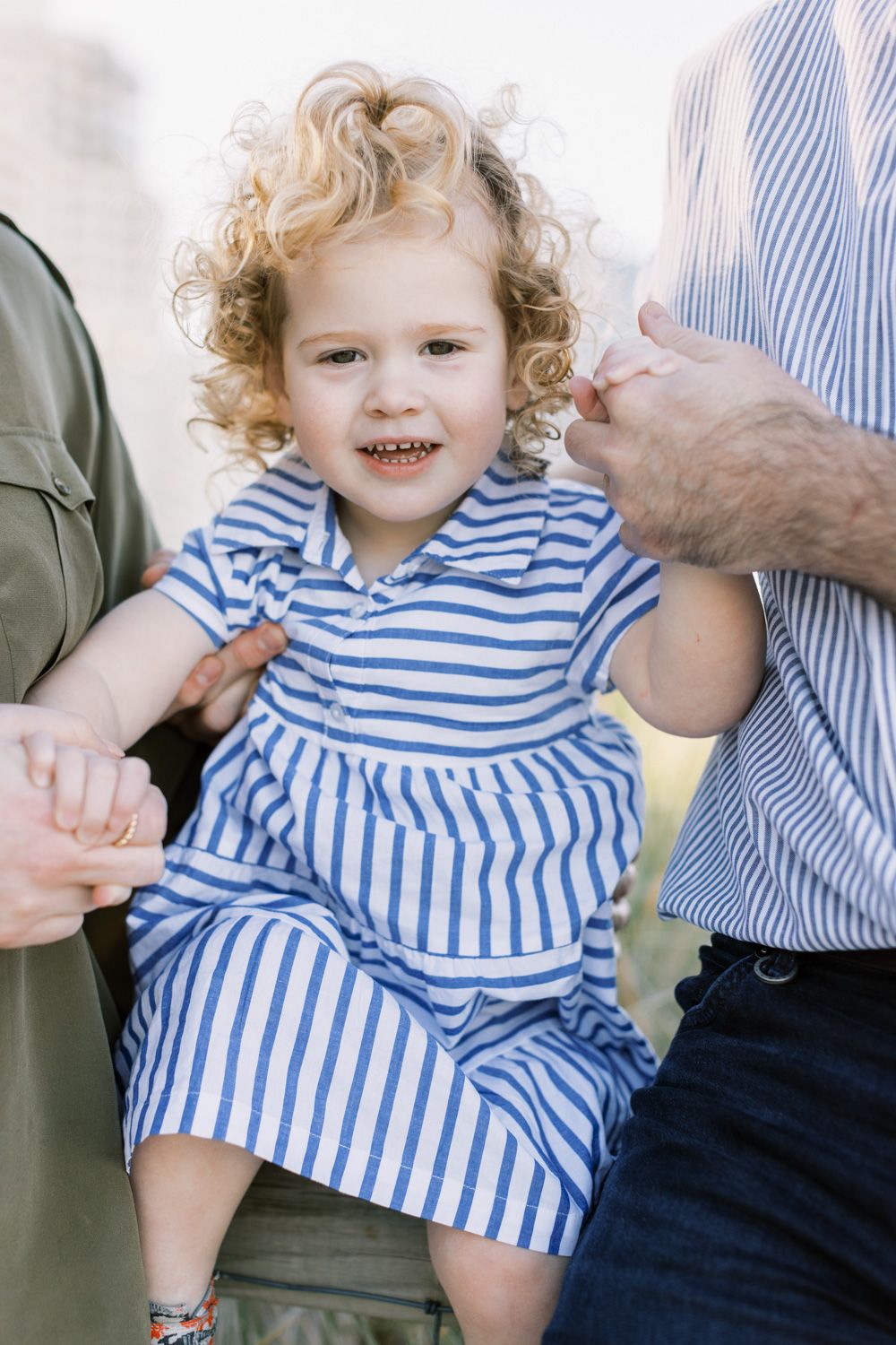 Child in blue striped dress with curly hair smiling while held by adults.