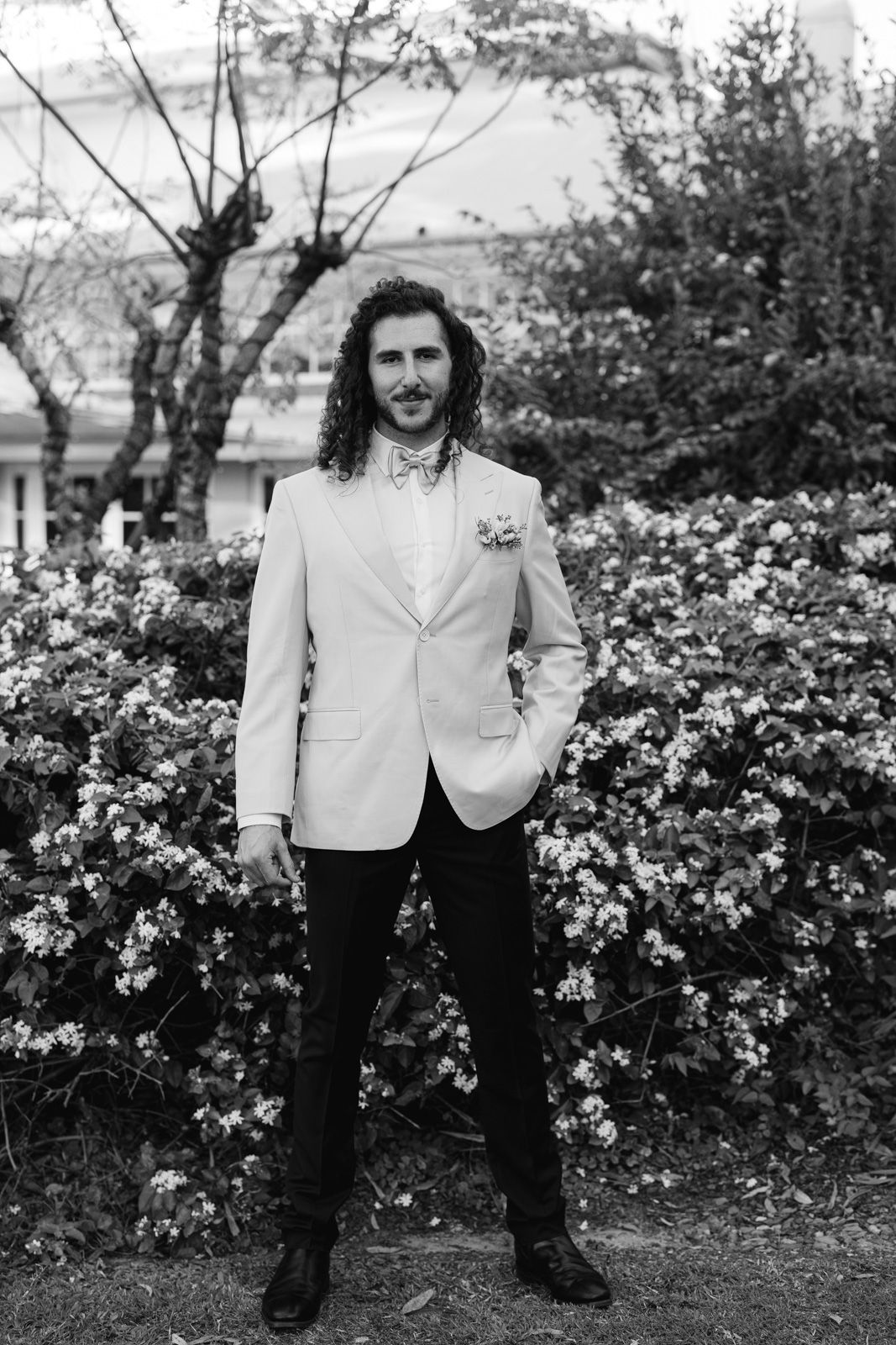 A person in a white blazer and black pants stands in front of blooming floral bushes in a black and white photograph.