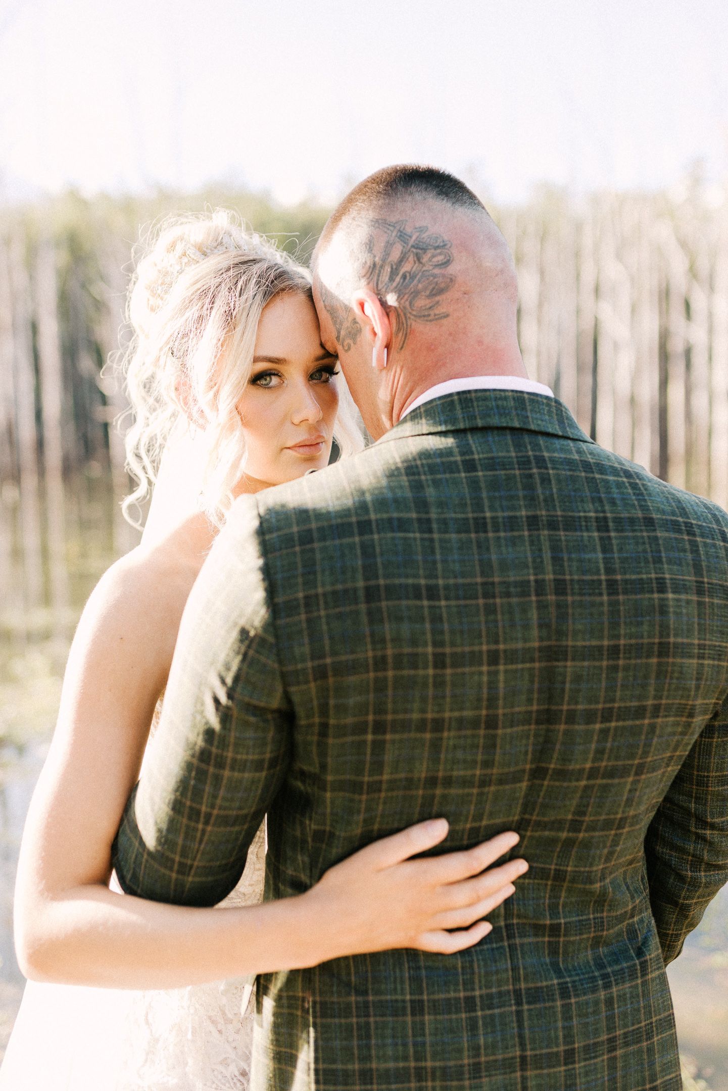 Bride in white lace gown embraces groom in green plaid suit, gazing over his shoulder at camera during outdoor wedding.