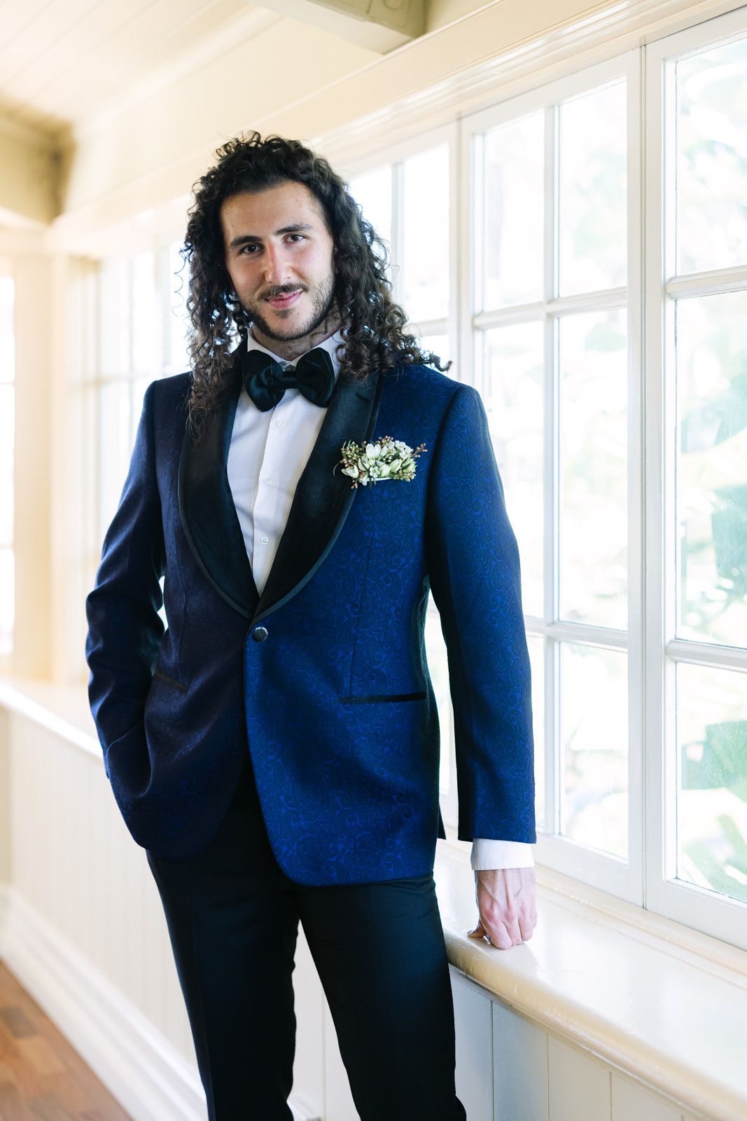 A groom in a navy blue tuxedo poses near bright windows in a wedding venue hallway.