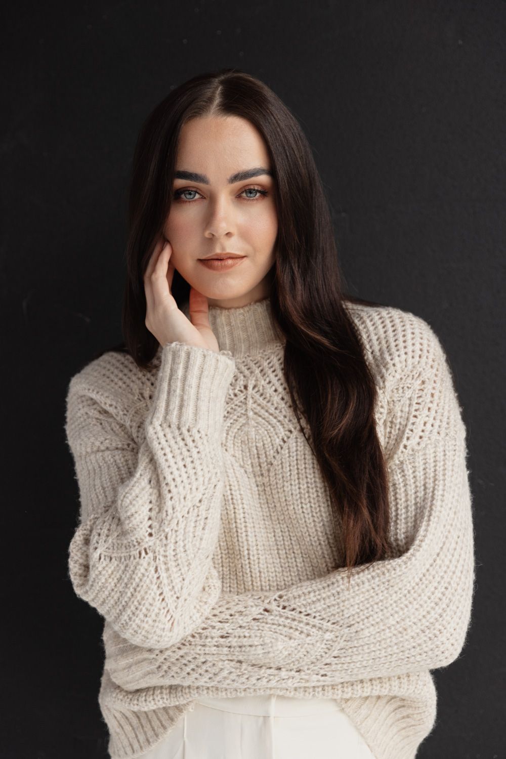 Portrait against dark background wearing cream knit sweater with thoughtful expression.