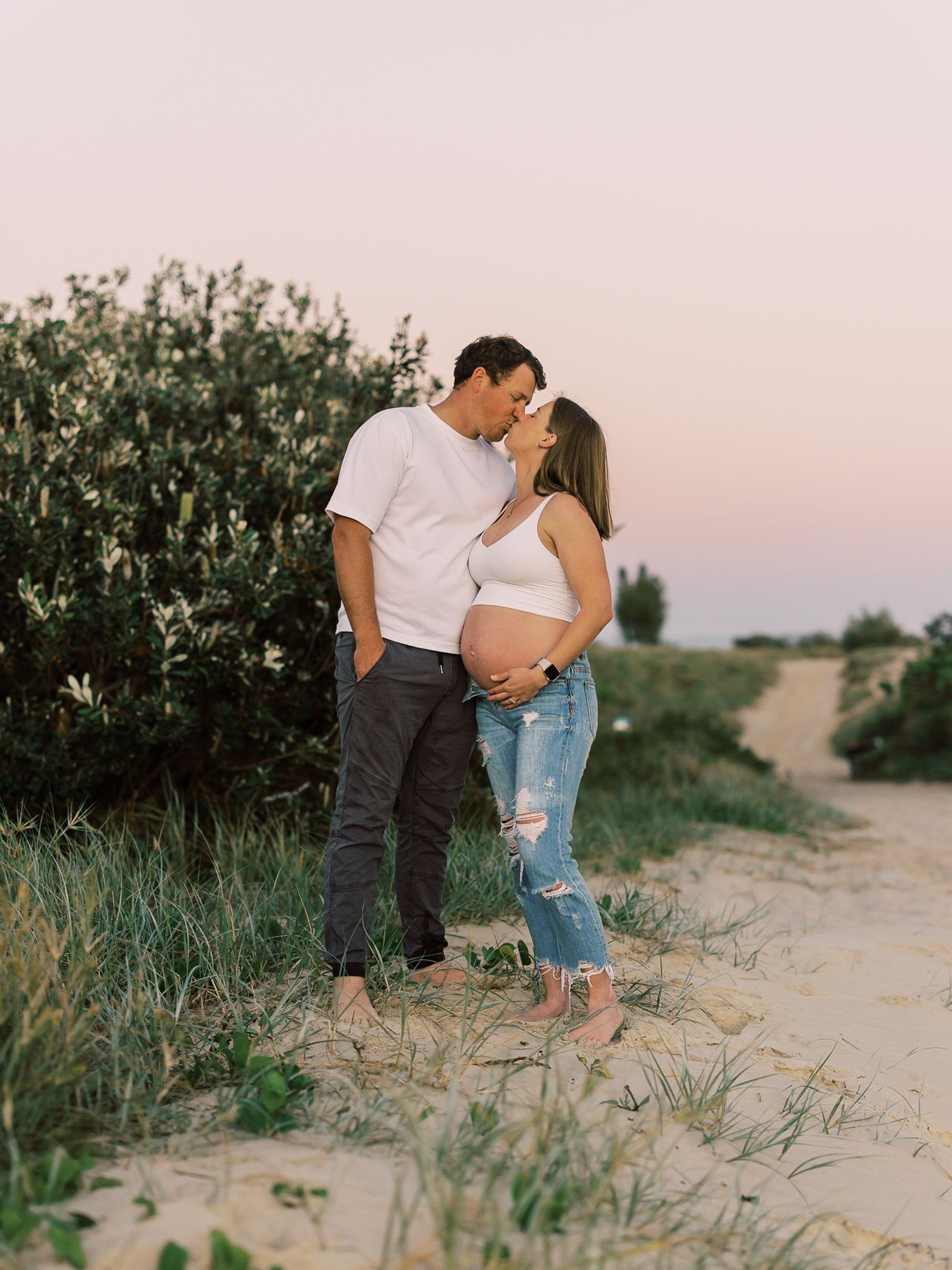 best maternity photo locations Gold CoastCouple embraces on sandy path during sunset with greenery and soft pink sky in background.