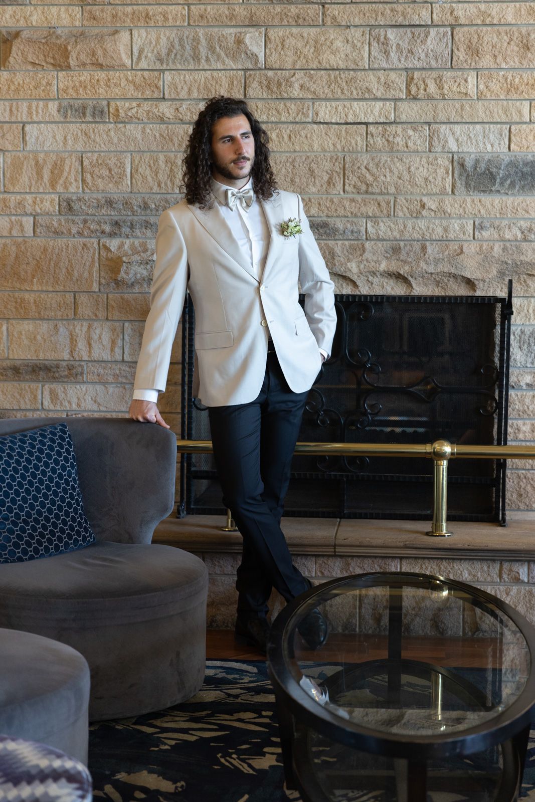A figure in a white blazer and dark pants poses near a stone wall and modern lounge furniture.