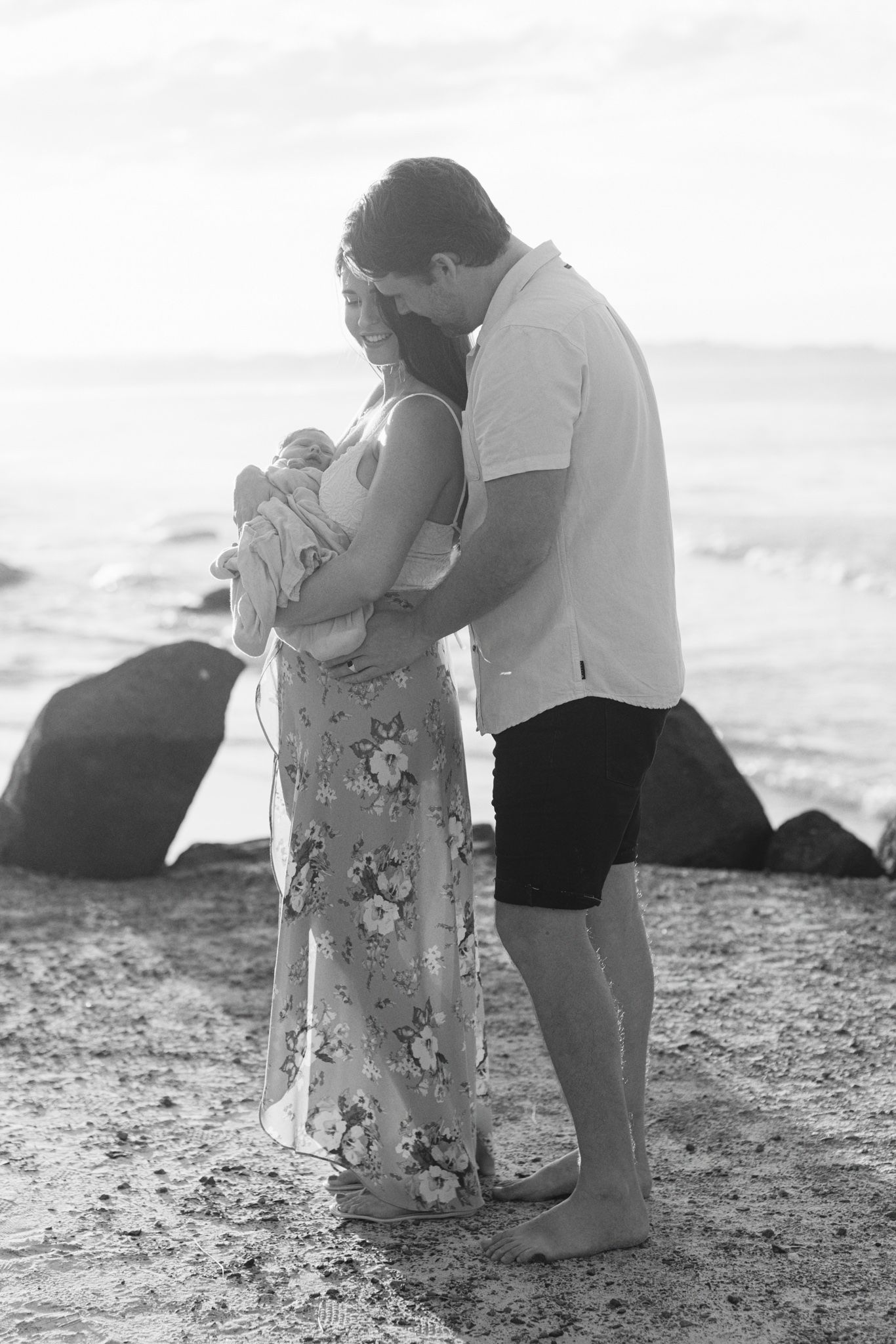 A couple stands together on a beach at sunset, with the woman wearing a floral dress and the man in casual attire.