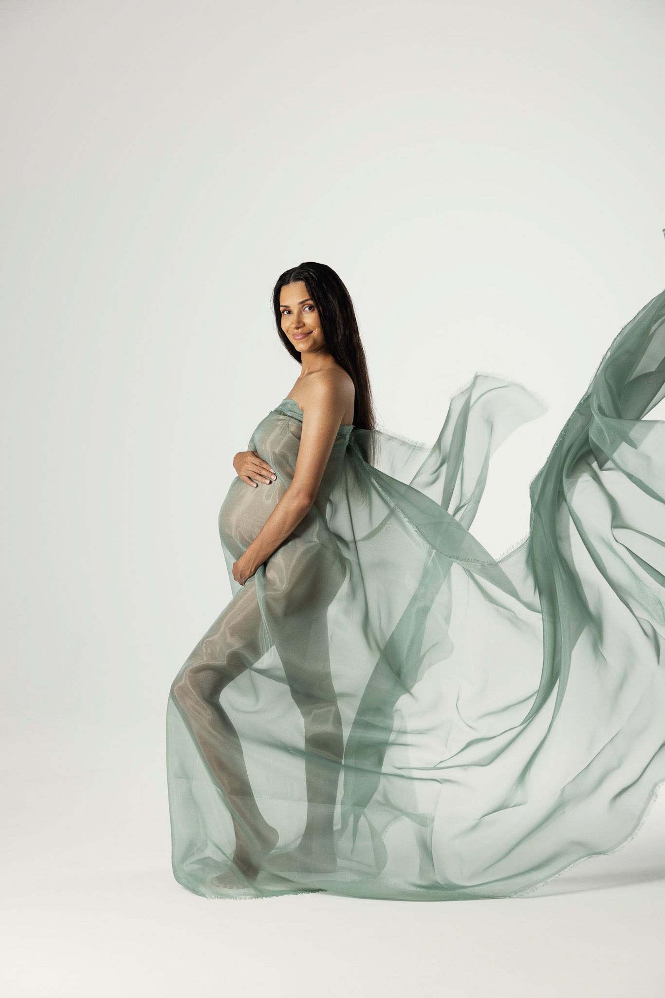 Ethereal maternity photo with flowing mint green fabric creating dramatic movement against white background.