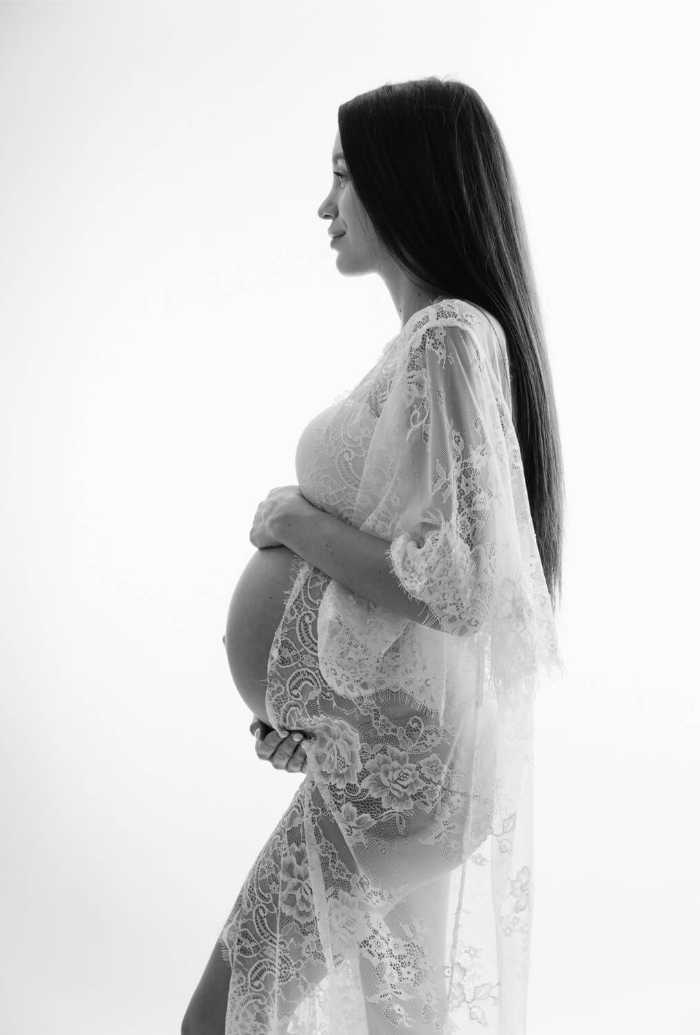 Black and white maternity photo of a pregnant woman in a lace robe cradling her baby bump against a white background.