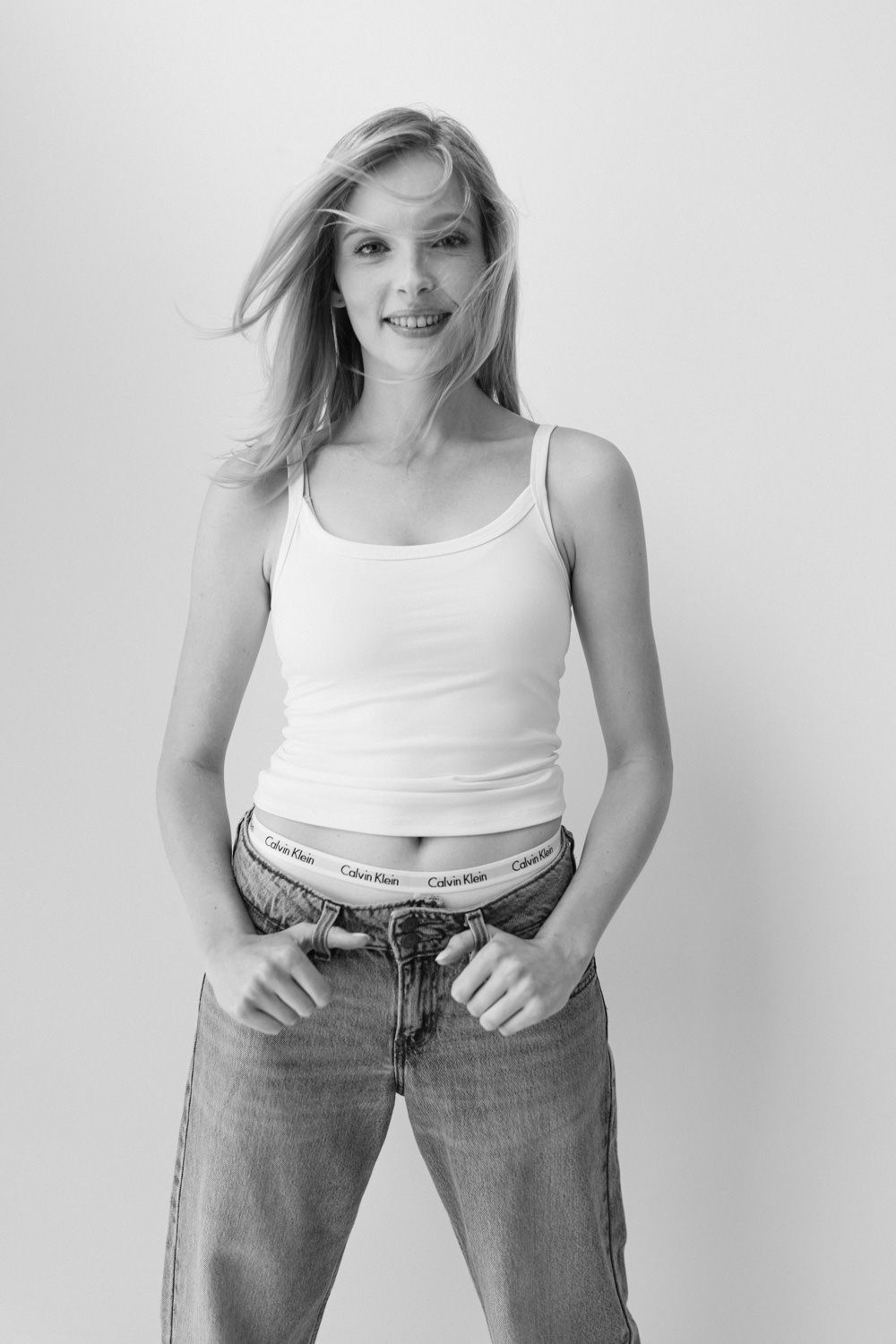 Black and white photo of blonde woman in white tank top and Calvin Klein jeans smiling.