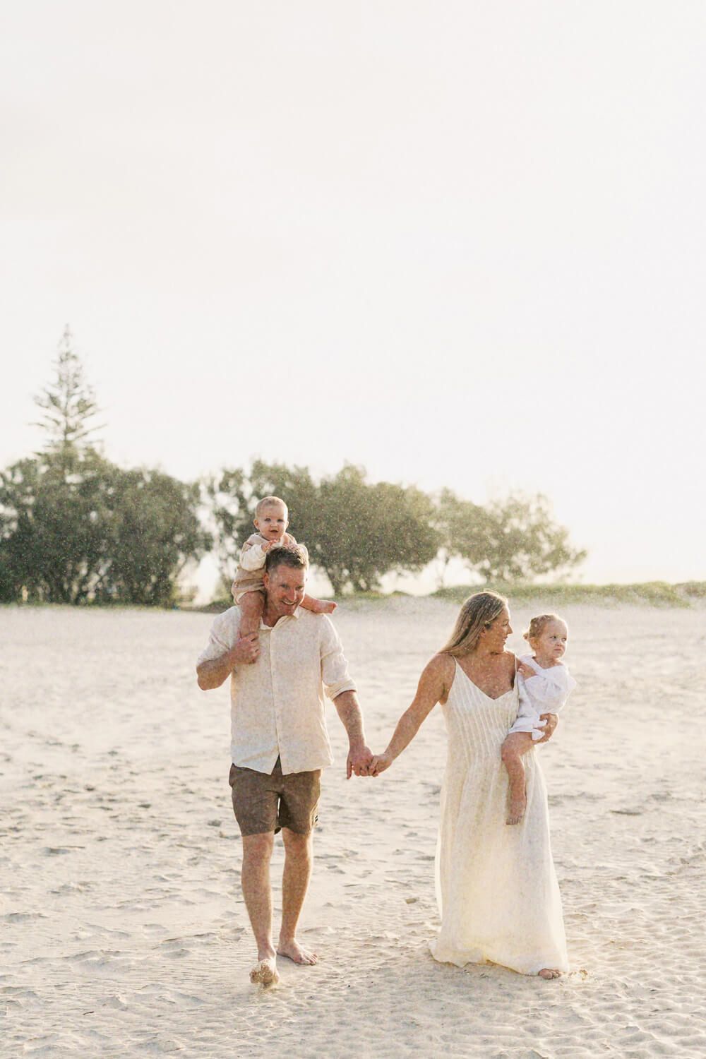 Happy family walking hand-in-hand on a sunny beach, dad with toddler on shoulders, mom carrying baby in white dress.