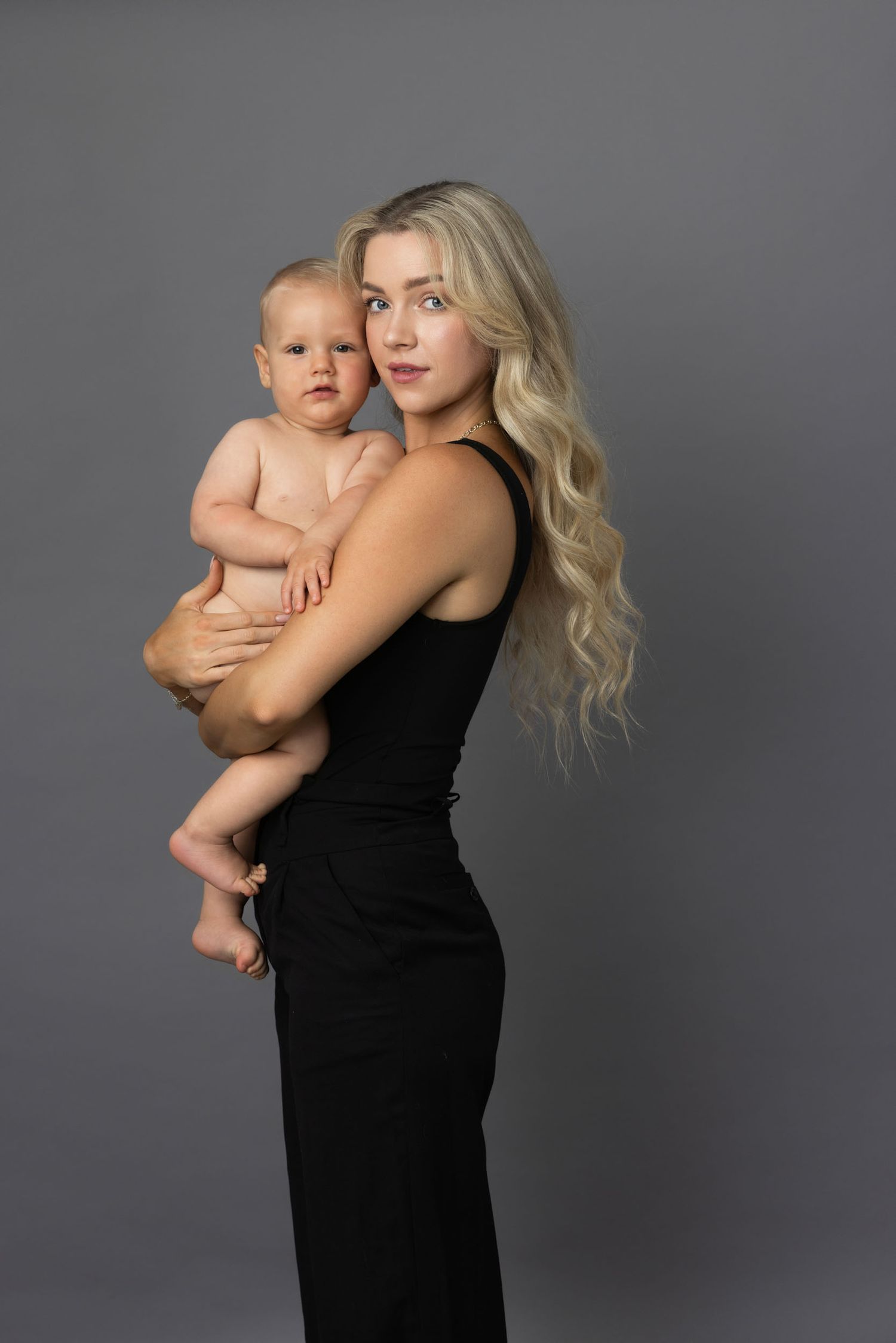 Studio portrait of a woman in a black dress holding a baby against a gray background.