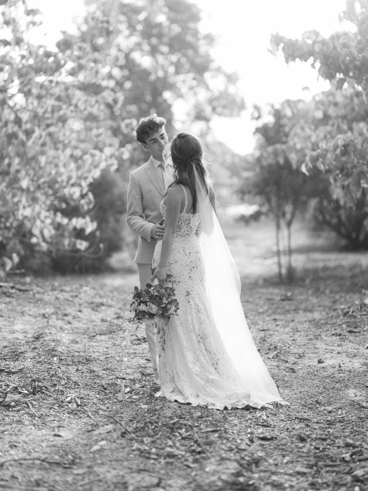 A romantic black and white wedding photo of a couple embracing on a tree-lined path with the bride holding a bouquet.