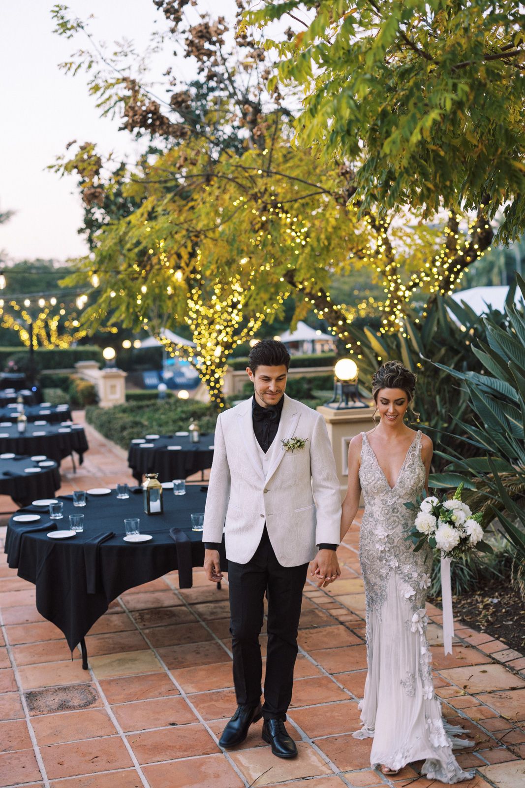 Elegant outdoor wedding reception setup with twinkle-lit trees, black tablecloths and terracotta tile patio.