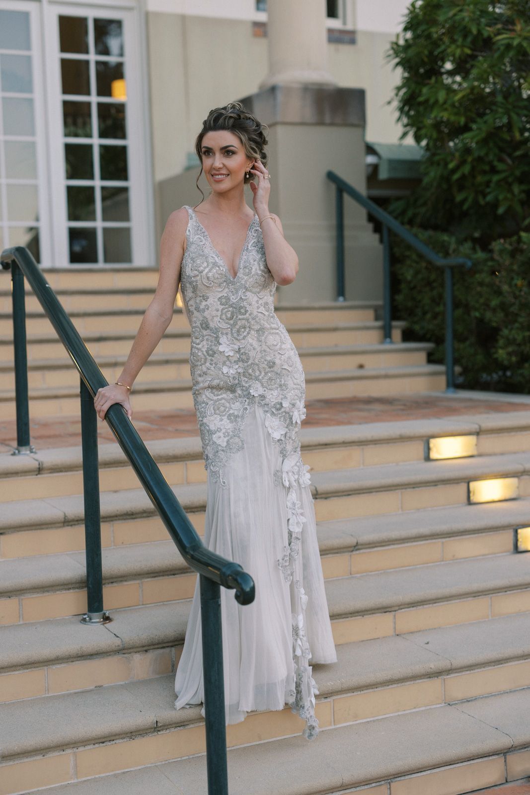 Elegant beaded wedding gown with plunging neckline and fitted silhouette photographed on outdoor stone steps at dusk.