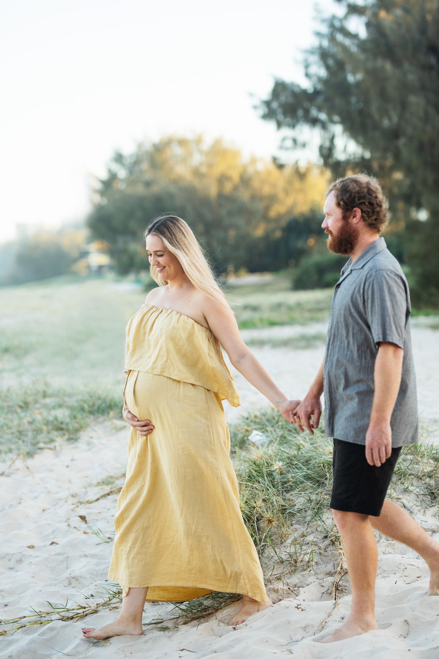 Romantic maternity photos of couple playing on sandy beach in yellow dress and casual wear.