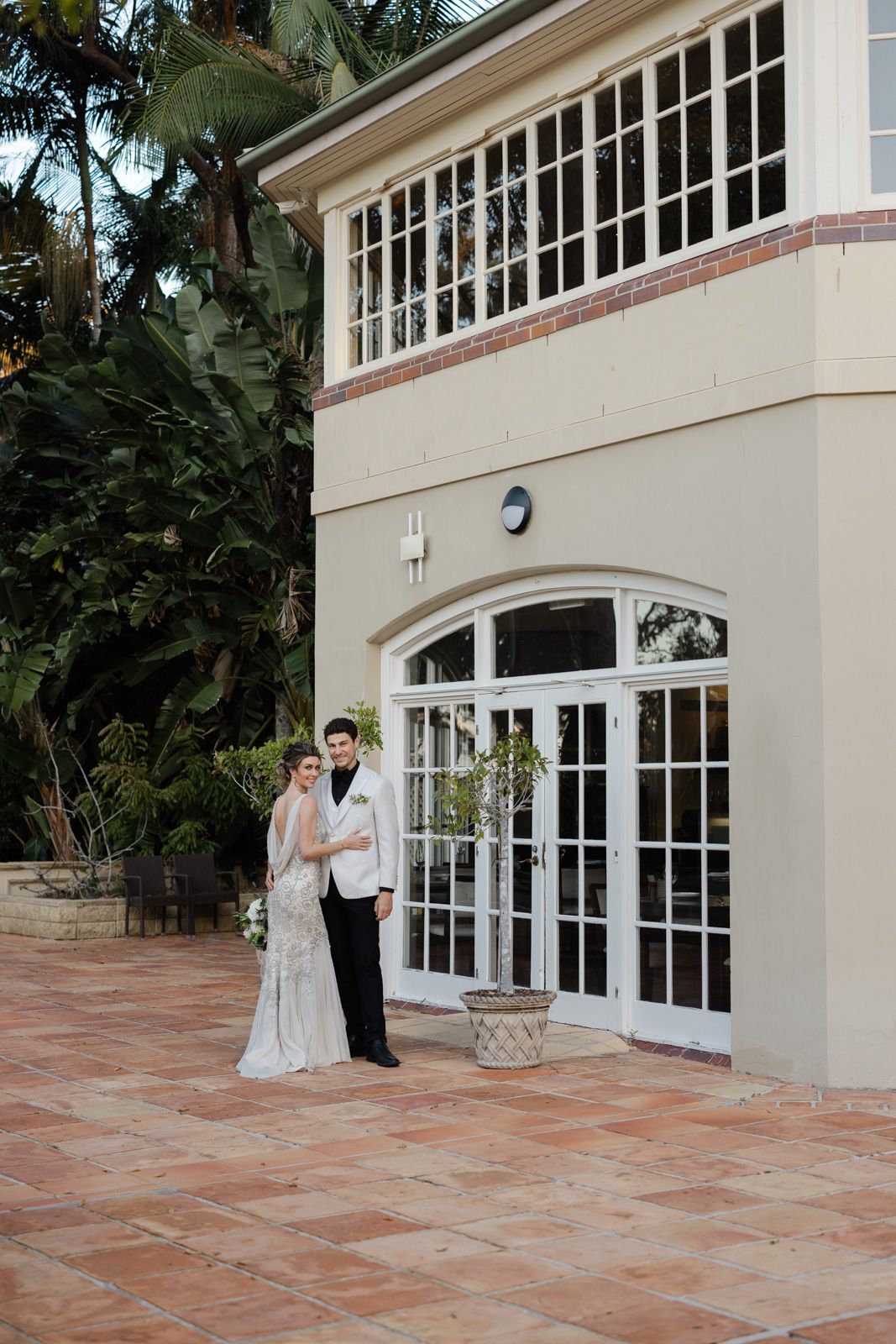 Elegant couple stands on terracotta tile patio outside cream-colored building with French doors and large windows.