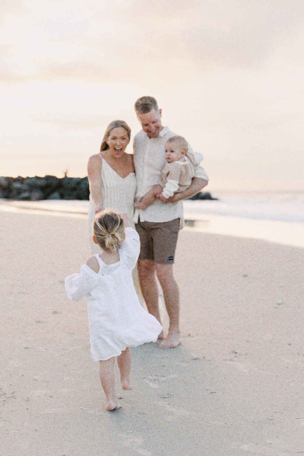 A joyful family watches their young daughter run toward them on a beautiful beach at sunset.