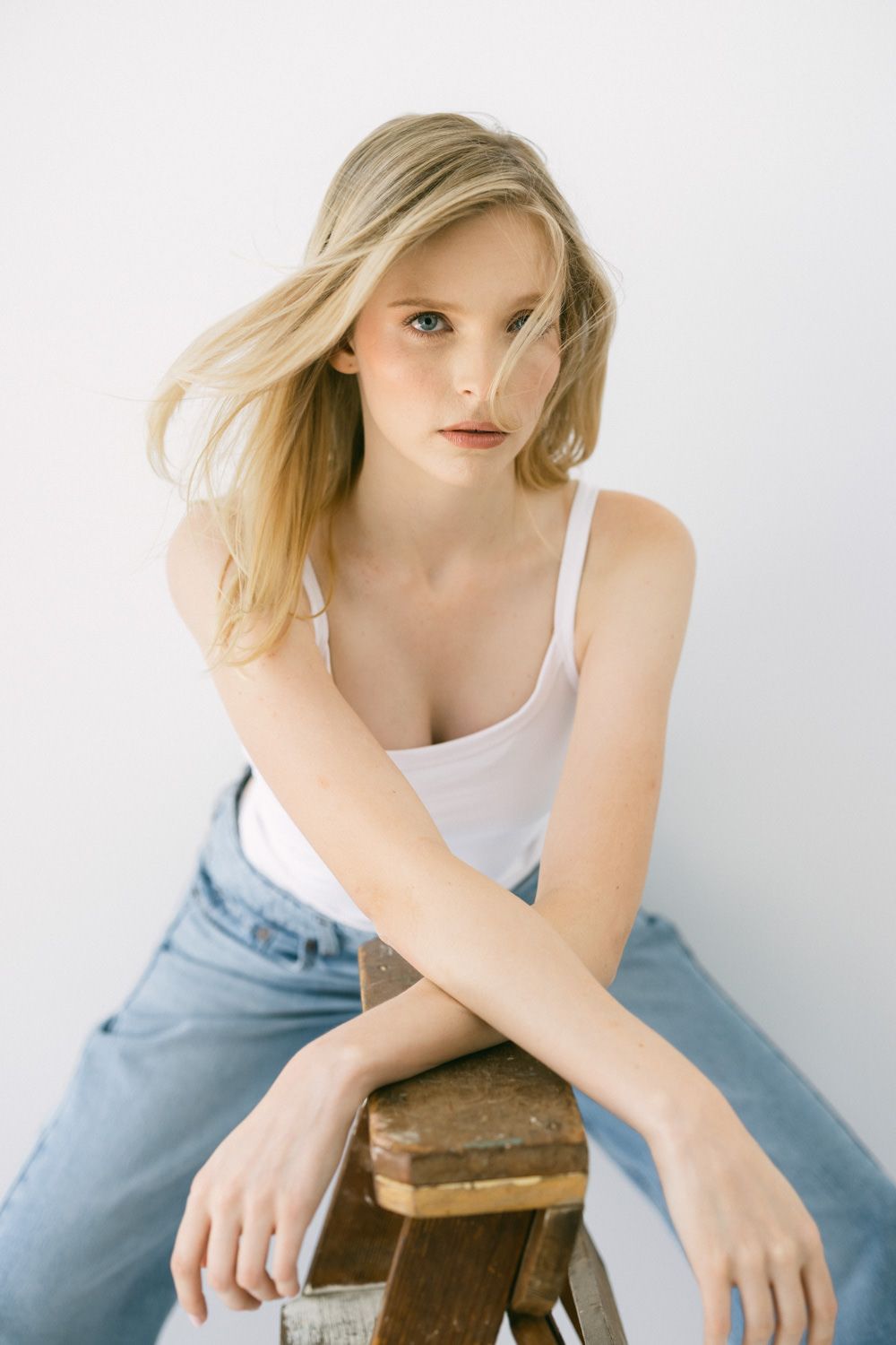 Young blonde woman in white tank top and jeans sitting on wooden stool against white background.