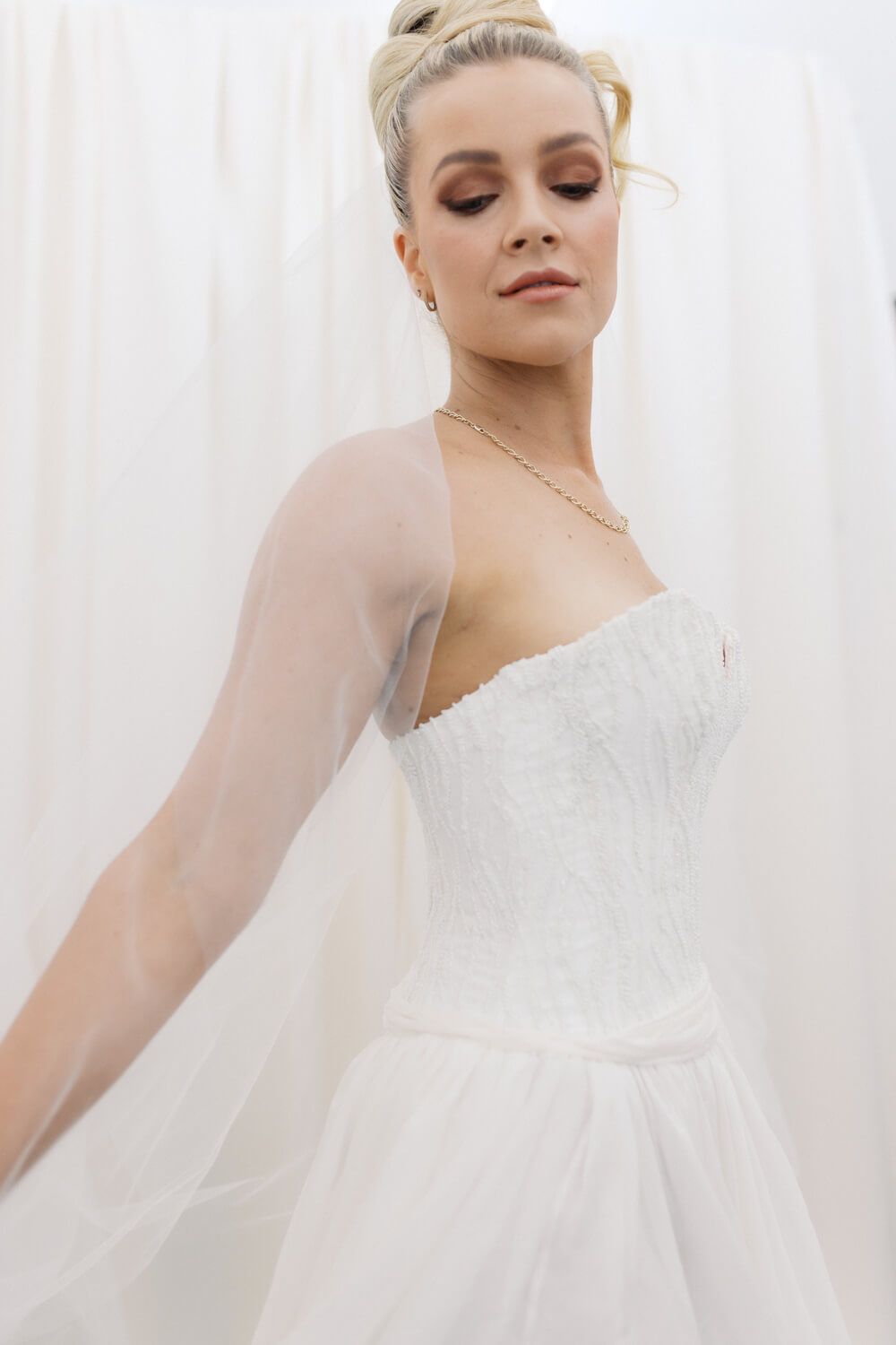 Elegant bride in a strapless white ballgown with sheer sleeves and lace bodice, hair up, soft makeup.