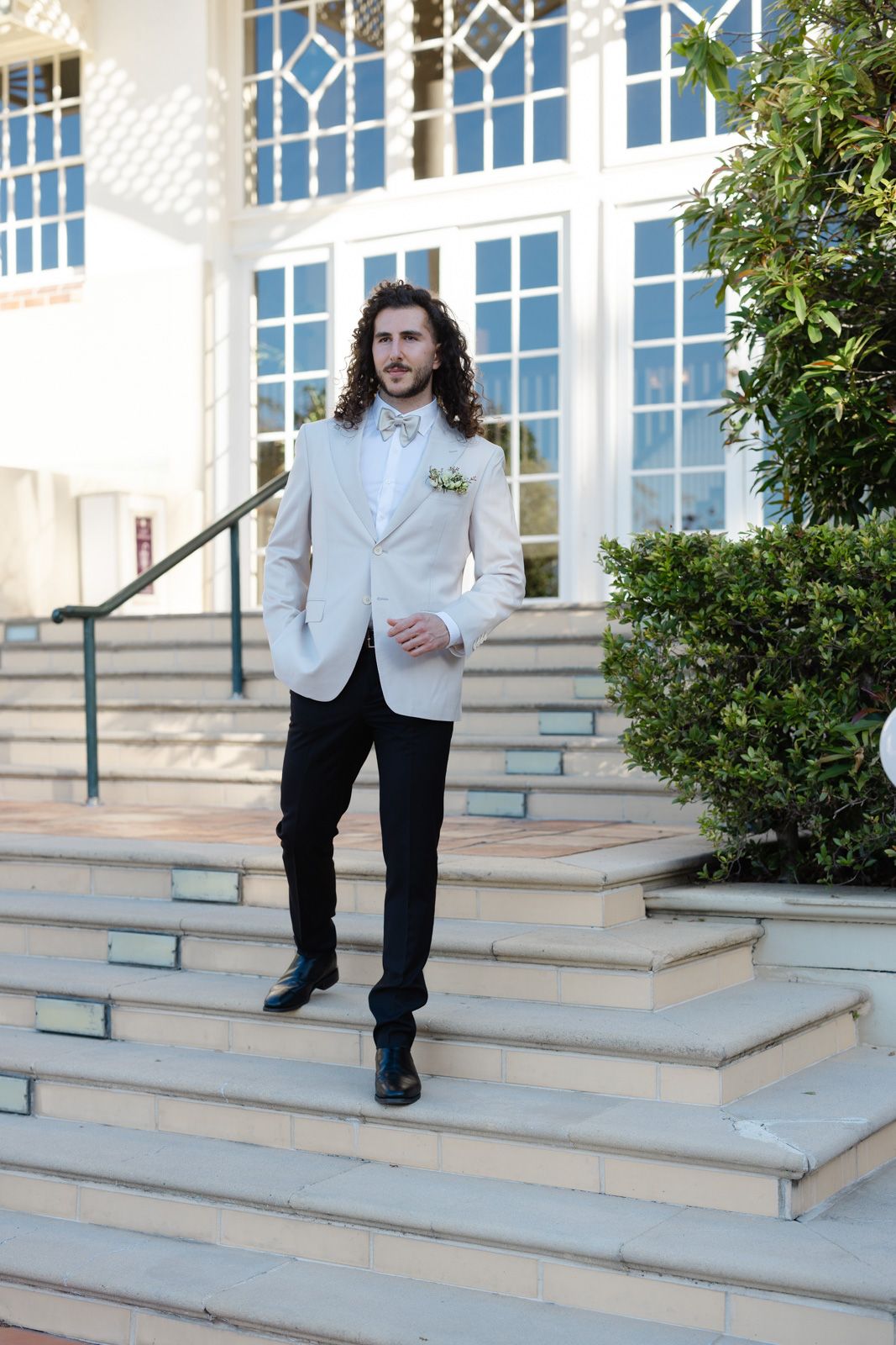 A person in a white dinner jacket and black pants stands on stone steps outside a building with ornate windows.