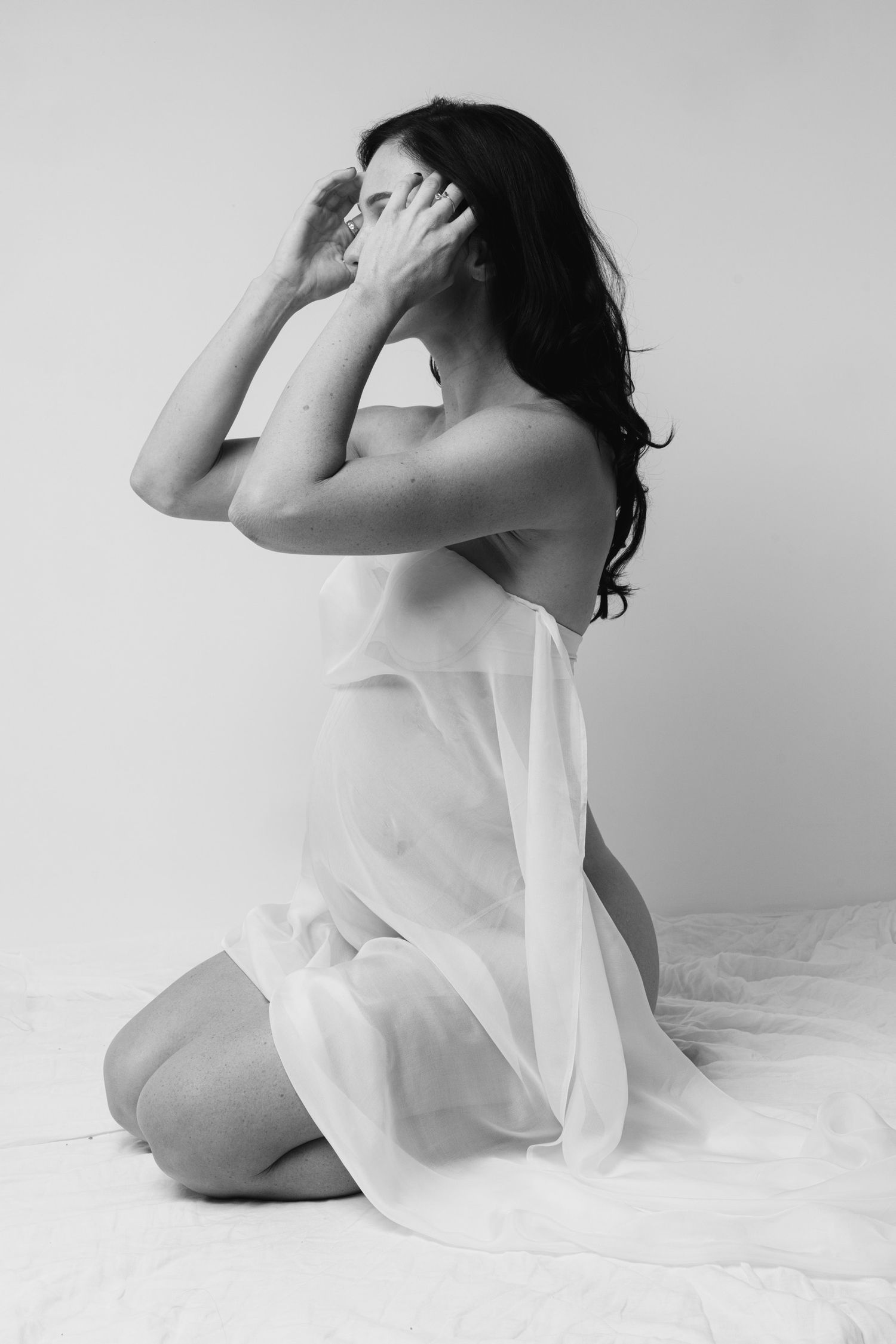 Artistic black and white portrait of a woman in flowing white fabric kneeling against light background.