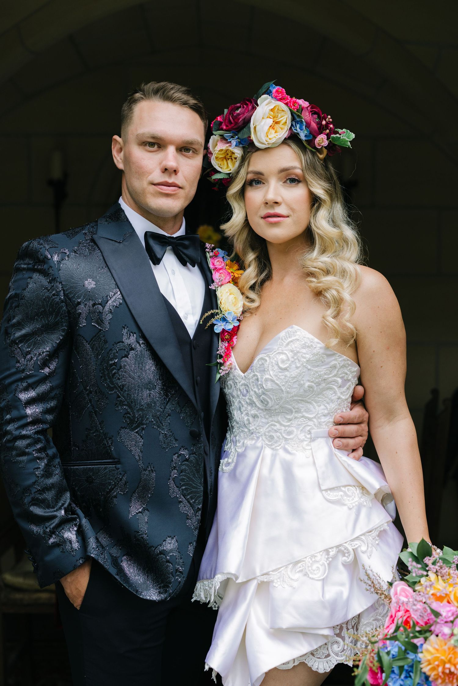 A wedding couple poses together in formal attire with the bride wearing a white dress and floral crown.