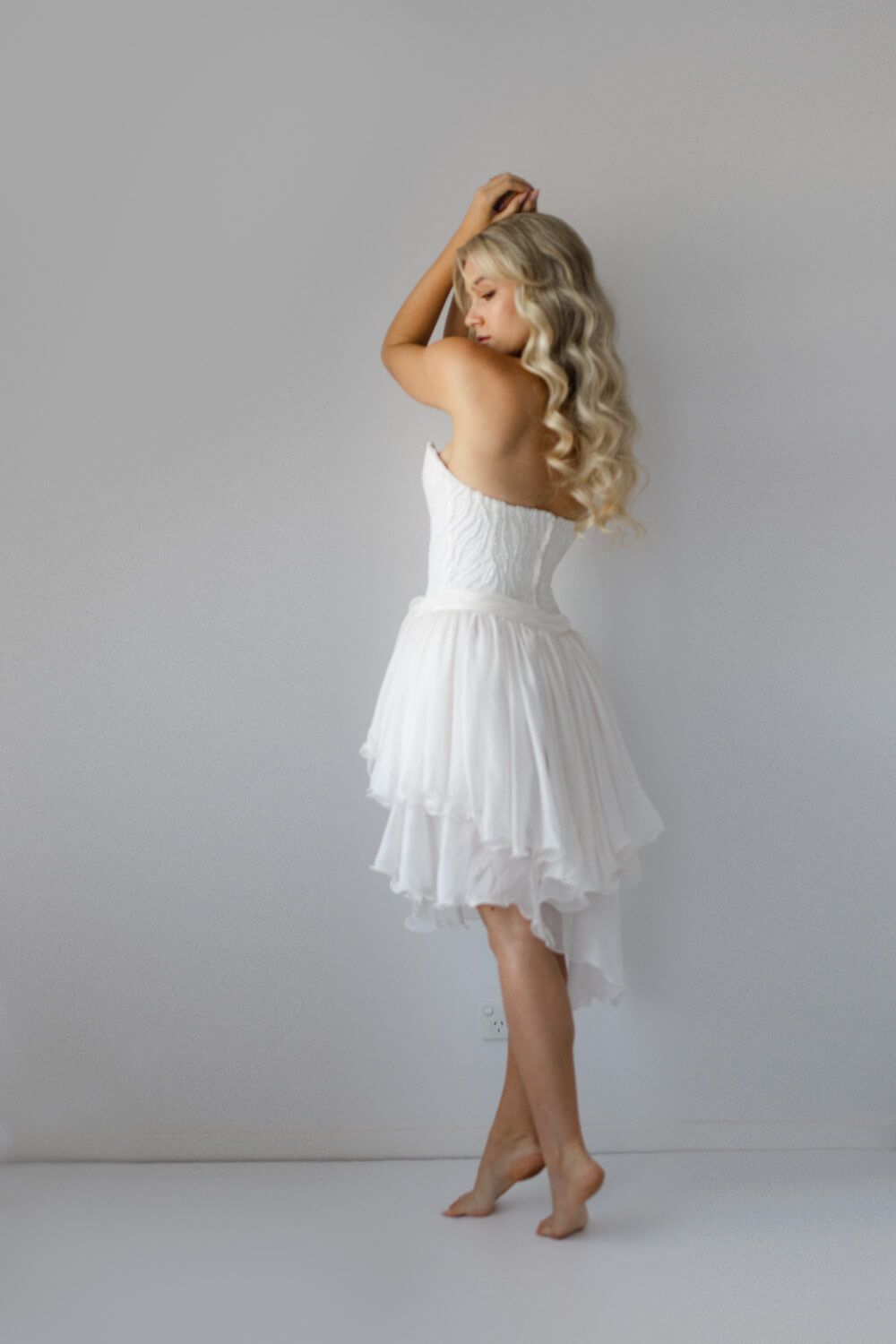 Barefoot blonde woman in a white strapless layered chiffon dress poses gracefully against a light gray background.