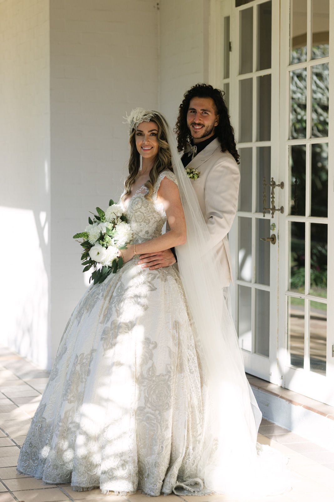 A bride and groom pose together on their wedding day wearing white attire with the bride holding a floral bouquet.