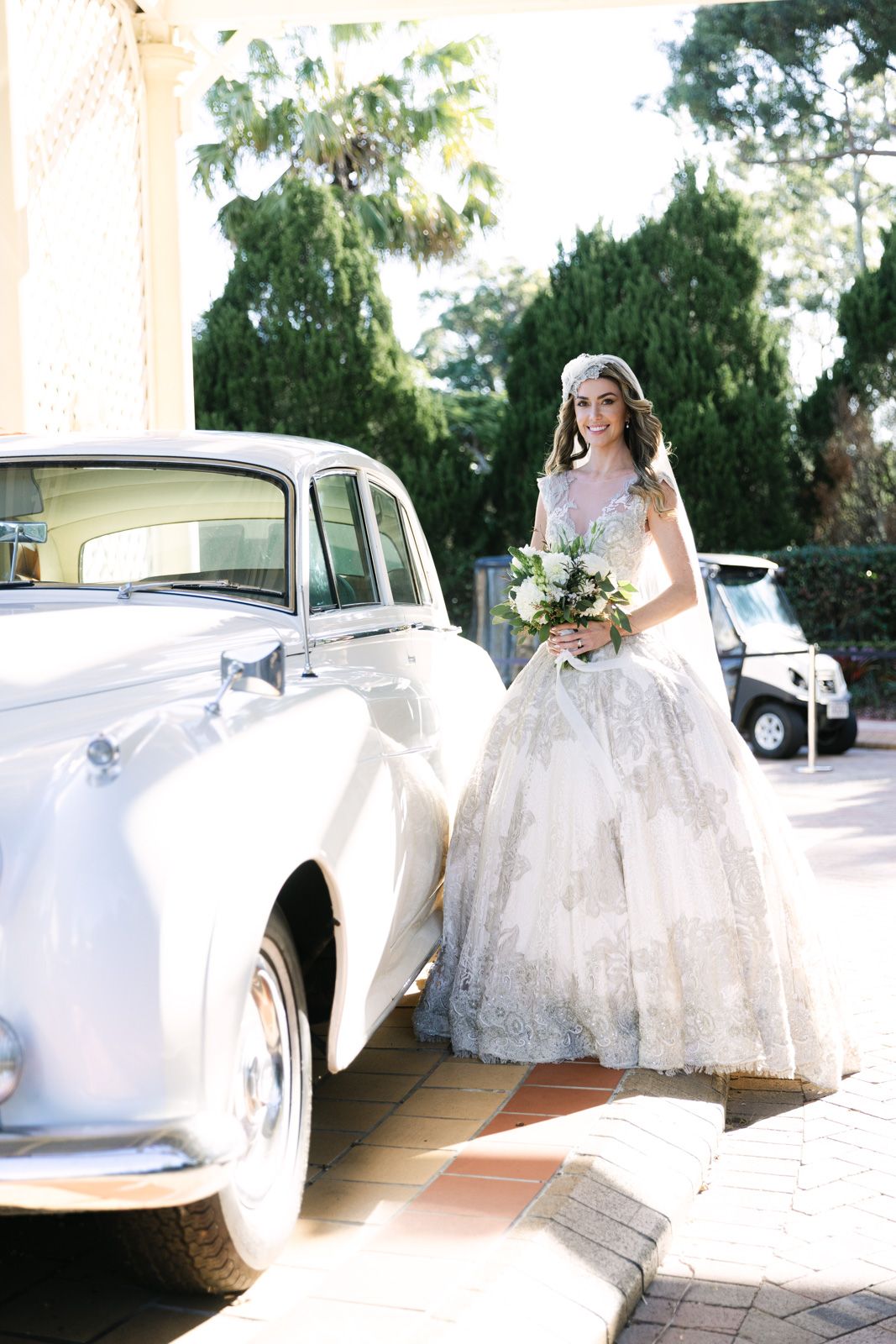 A classic white vintage car parked beside someone in an elegant lace wedding gown holding a floral bouquet.