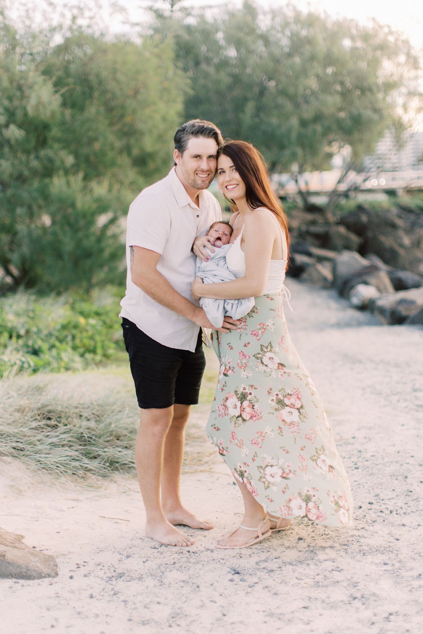 A family shares tender moments together on a sandy beach path surrounded by natural greenery and rocks.