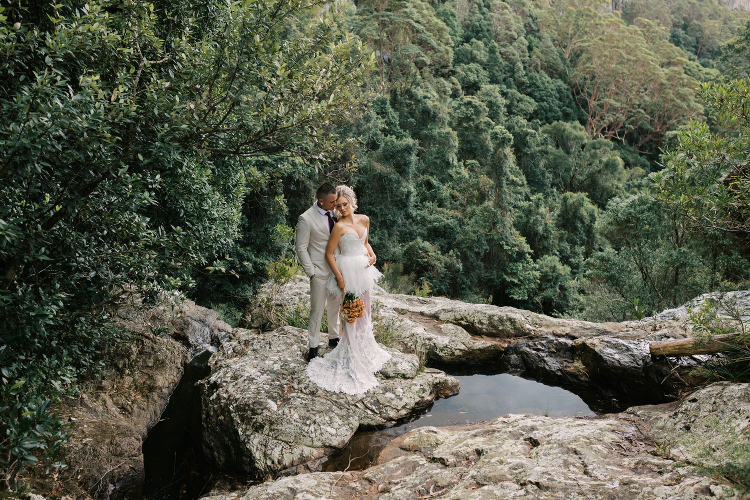 Bride and groom embrace on rocky outcrop surrounded by lush green forest in a romantic outdoor wedding photo.