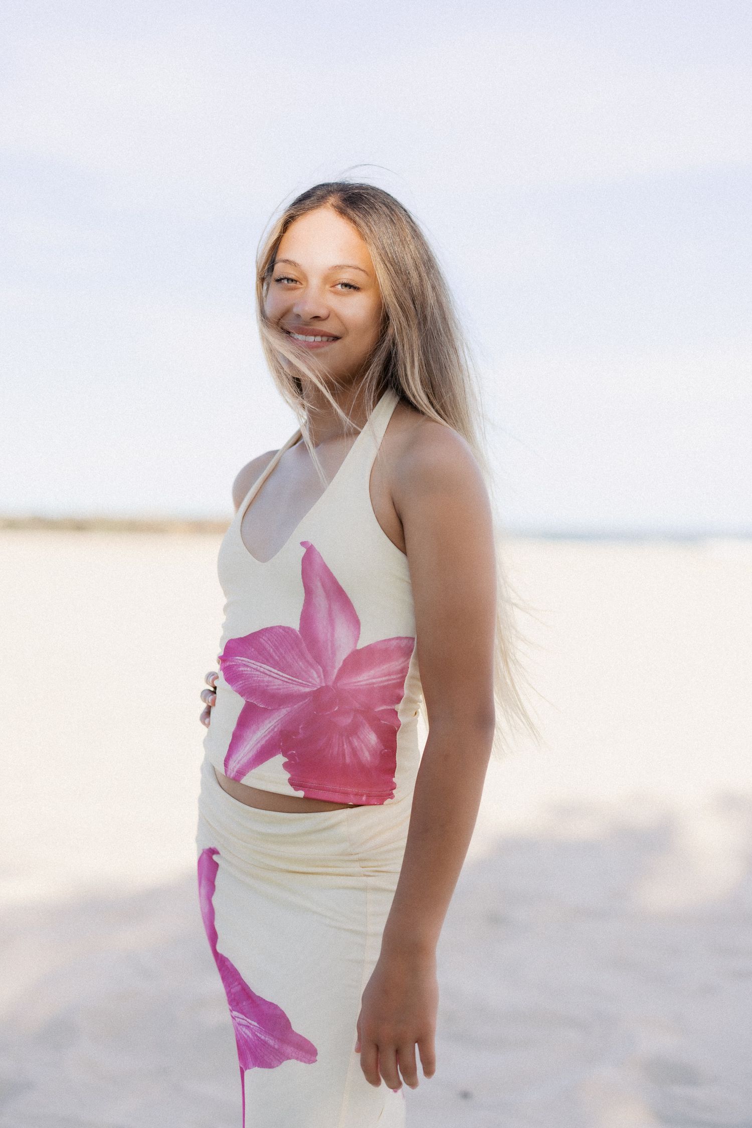 Smiling girl with long blonde hair wearing a floral crop top and skirt set on a sandy beach.