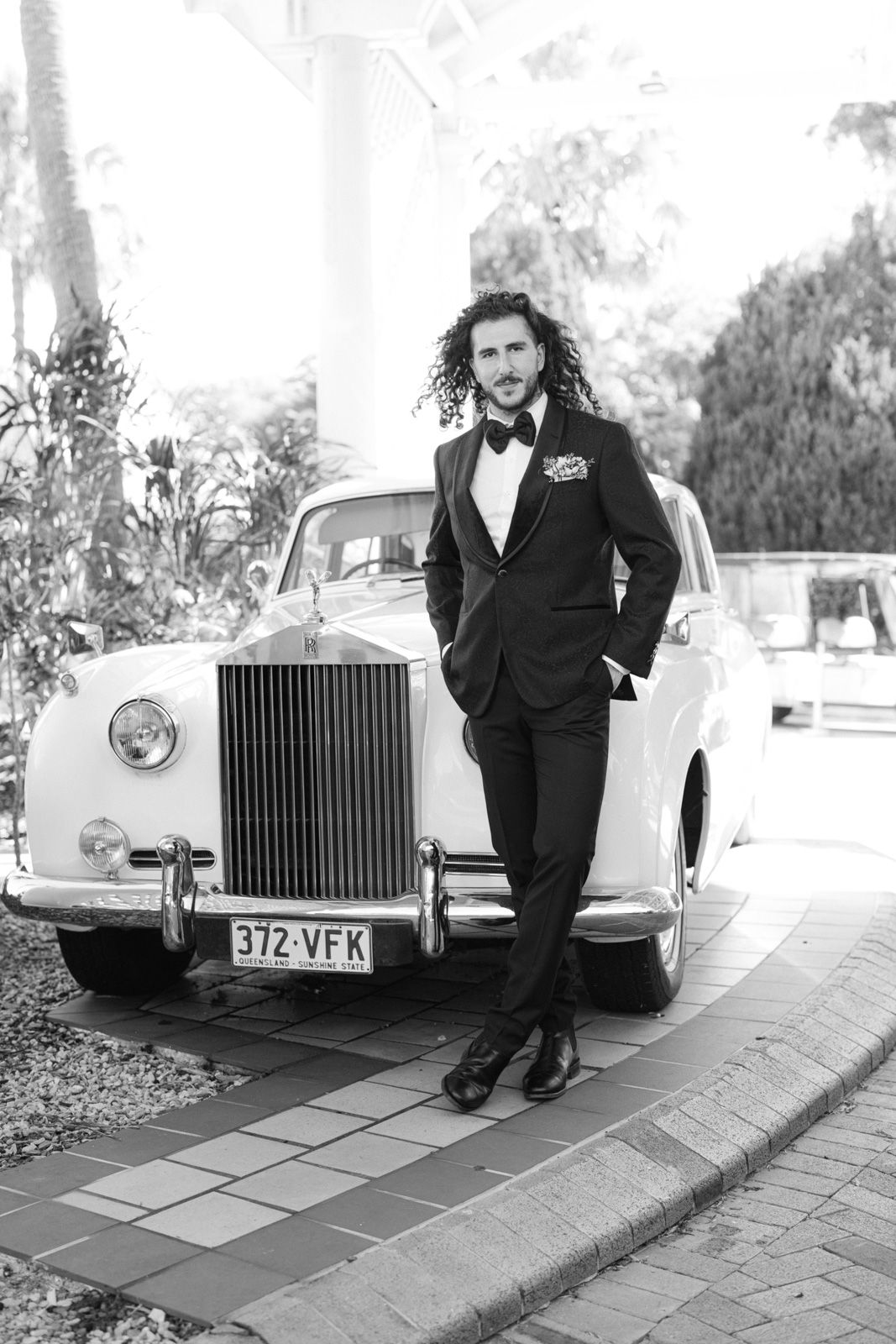 Classic white Rolls Royce luxury car parked on brick path with person in formal black tuxedo standing beside it.