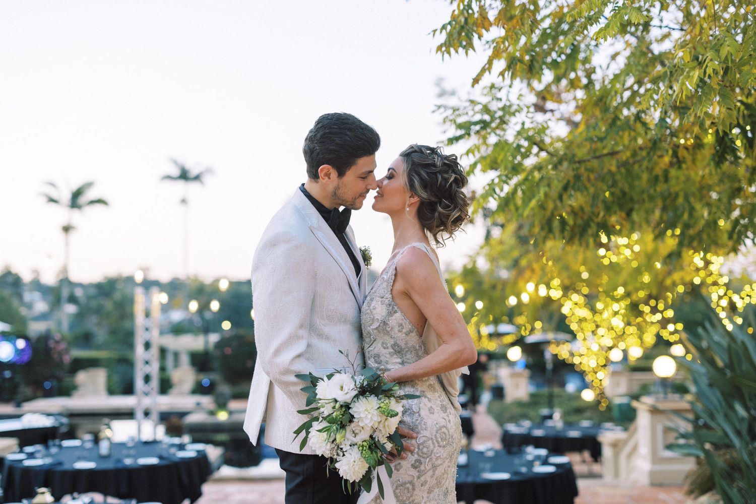 A romantic outdoor wedding scene with a couple kissing under golden sunlit trees and white floral bouquet.