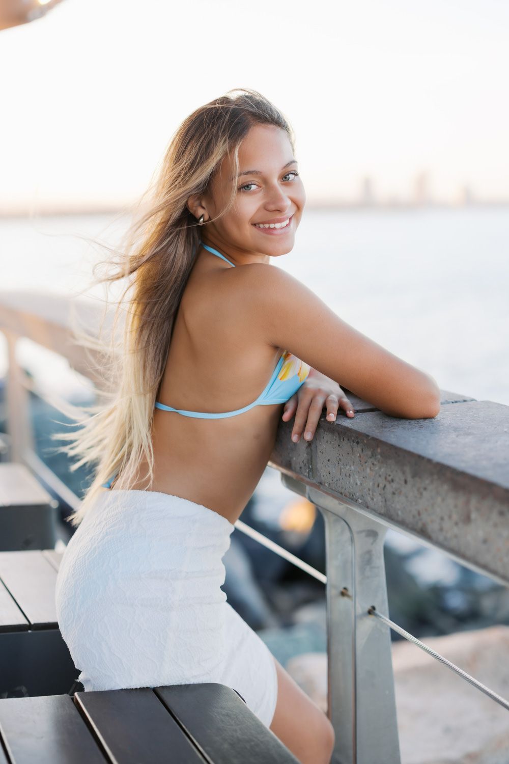 Young woman in blue bikini top and white skirt smiling on a waterfront pier at sunset.