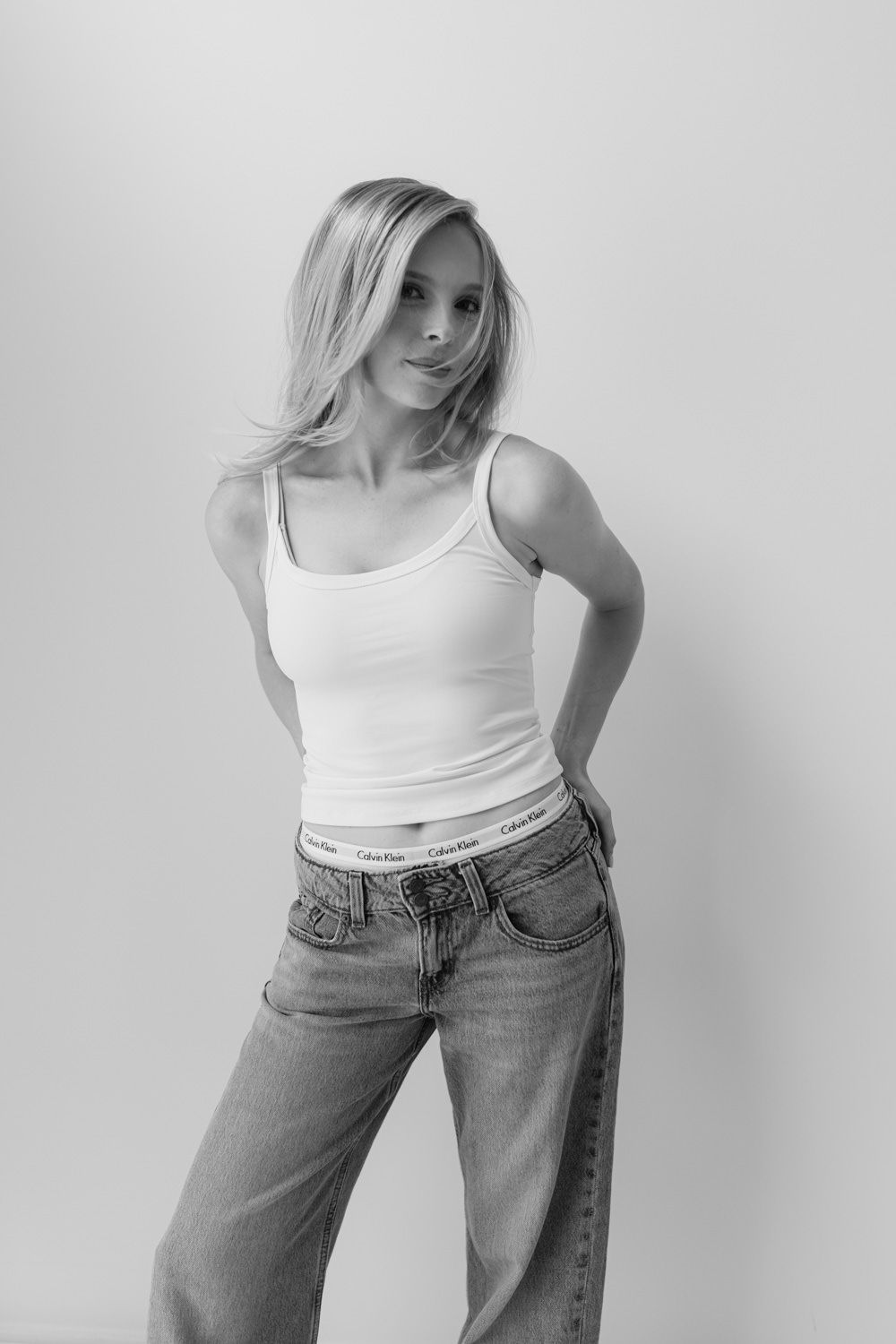 Black and white photo of a blonde woman in a white tank top and Calvin Klein jeans posing against a light background.