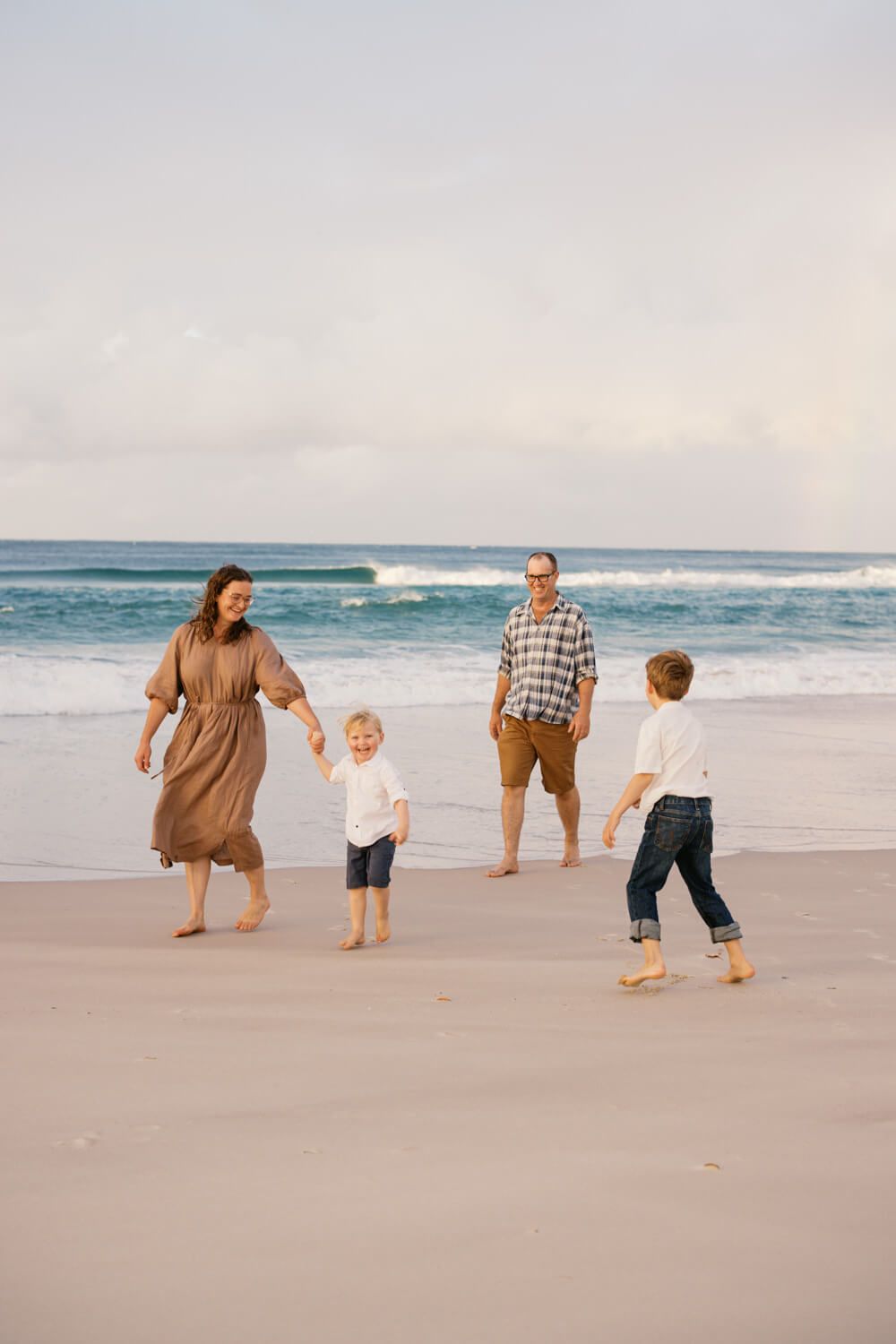 Happy family of four walking together on a sandy beach with ocean waves in the background at sunset.