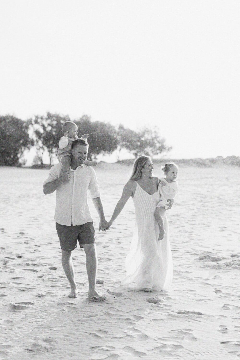 Black and white photo of a family walking on a sandy beach, parents holding hands while carrying young children.