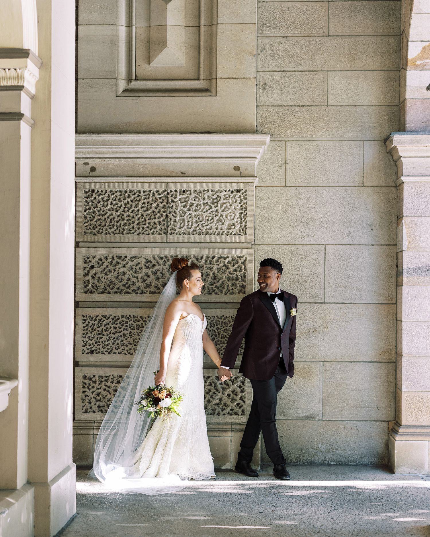 A wedding couple poses against an ornate stone building with architectural details.