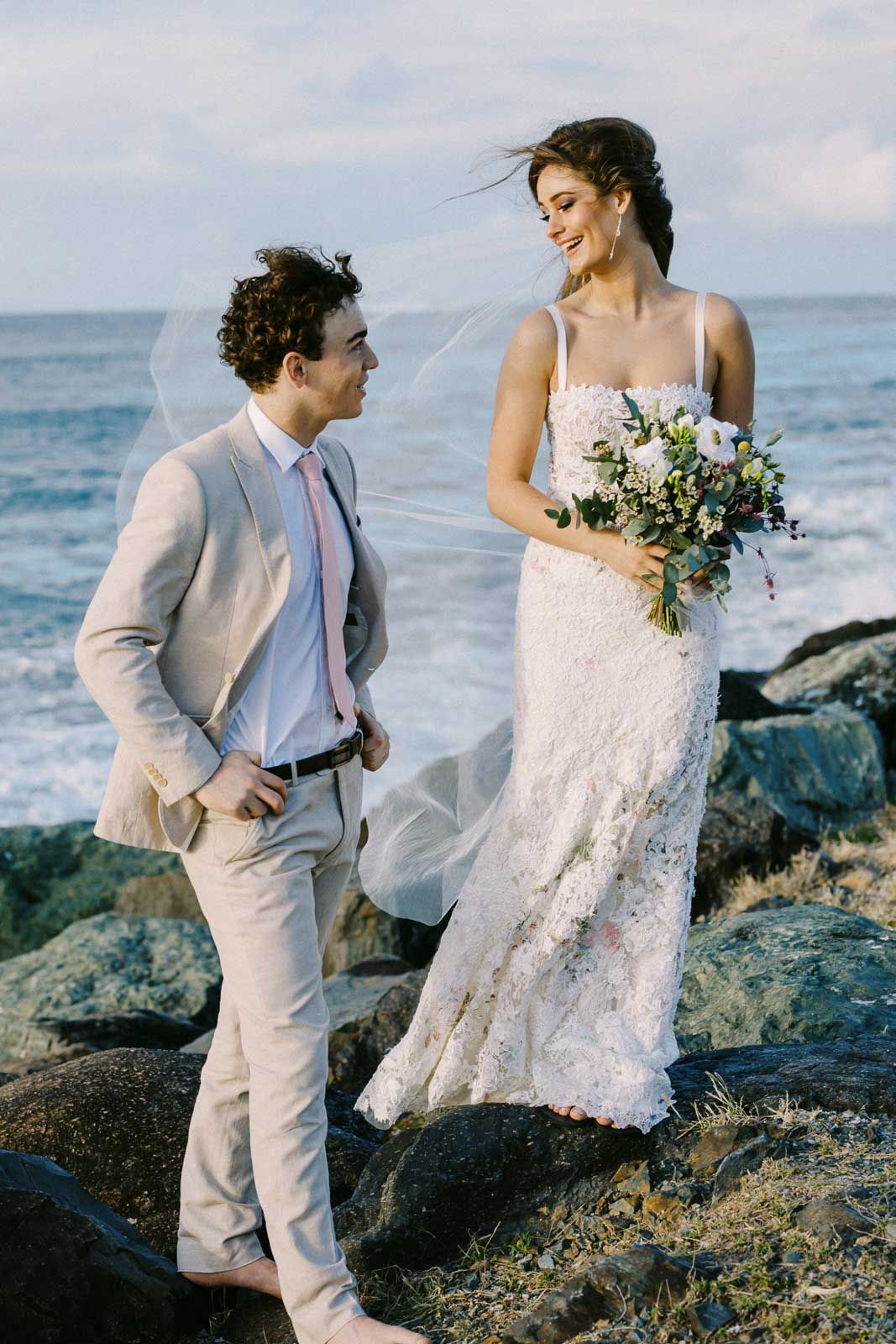 A romantic beach wedding scene with a couple in elegant attire standing on coastal rocks by the ocean.