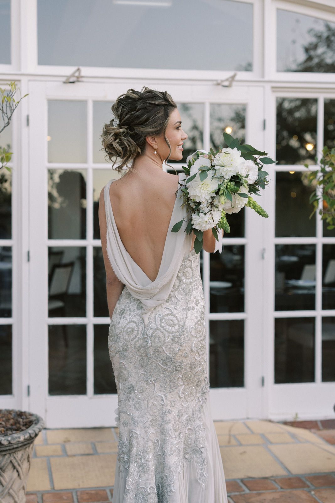 A bride in a white lace wedding gown with cowl back detail holds a bouquet of white flowers against French doors.
