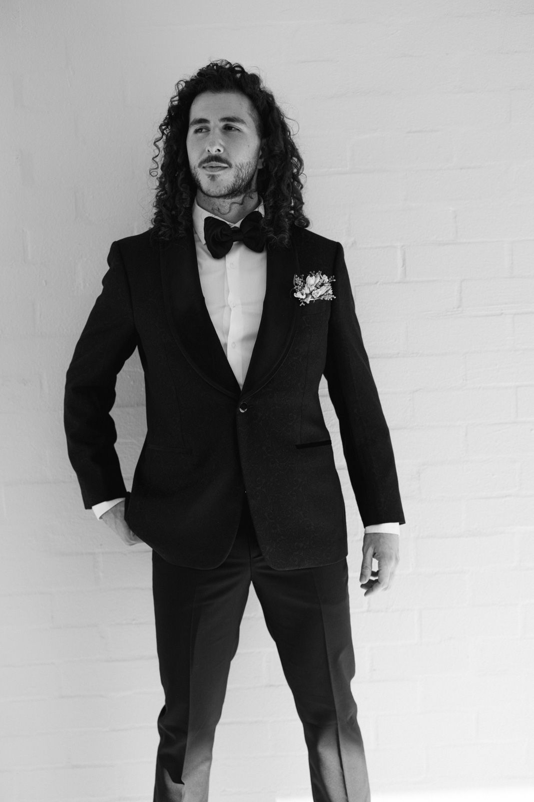 A black and white portrait of someone in a formal tuxedo with bow tie, curly hair, and boutonniere against a white wall.