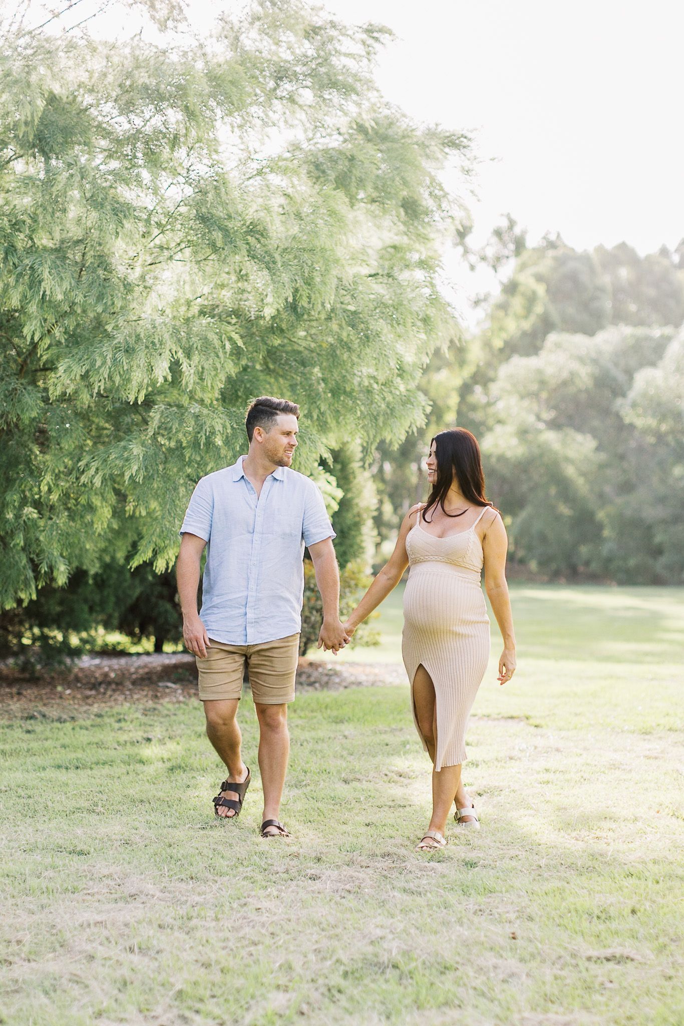 A couple walks together on a grassy field near evergreen trees during a sunny afternoon.