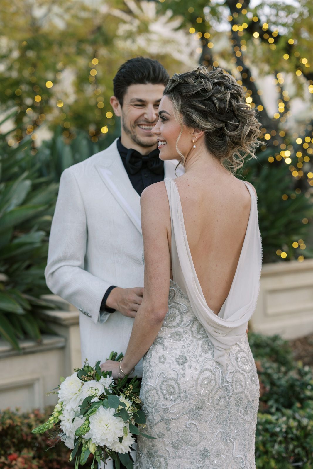 A romantic wedding couple shares intimate moments in a garden setting with twinkling lights and white floral decor.