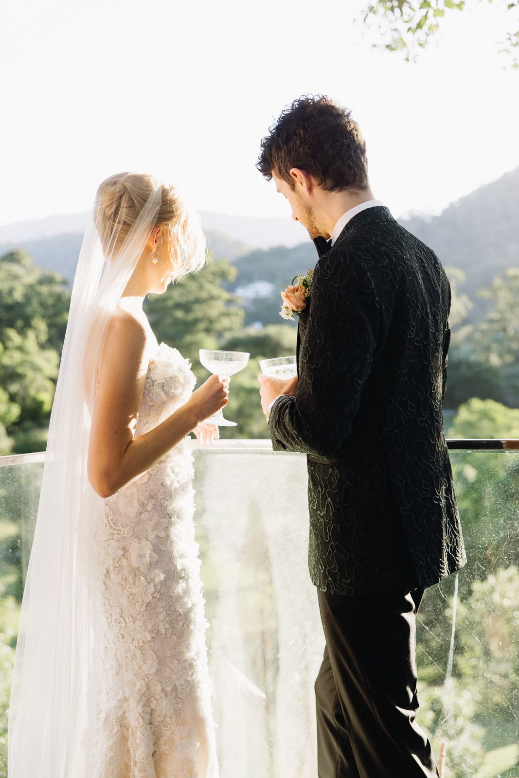 Newlywed couple shares a toast on a scenic balcony overlooking mountains during their wedding celebration.