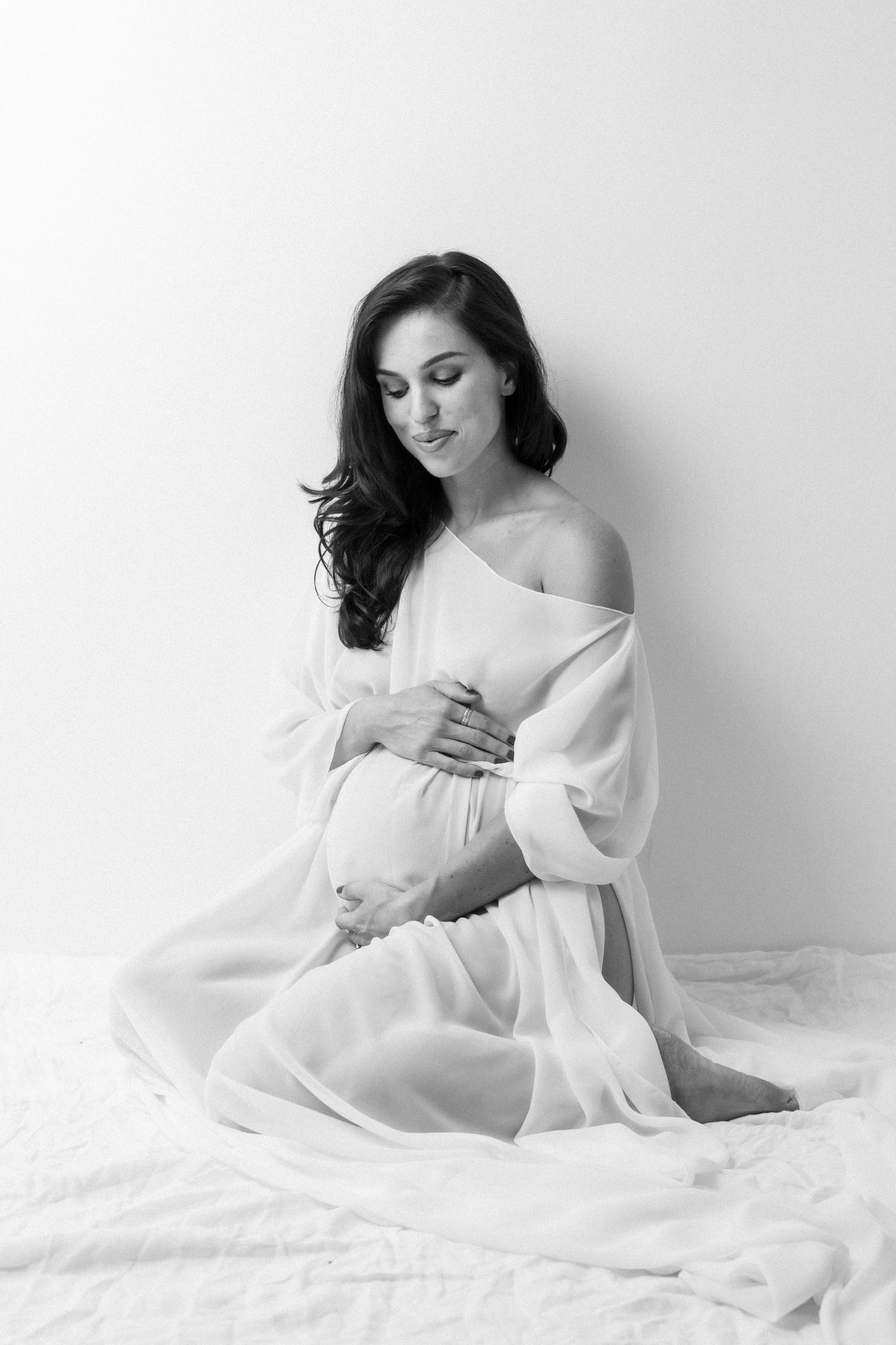 A black and white maternity portrait showing an elegant figure in a flowing white dress sitting on white bedding.