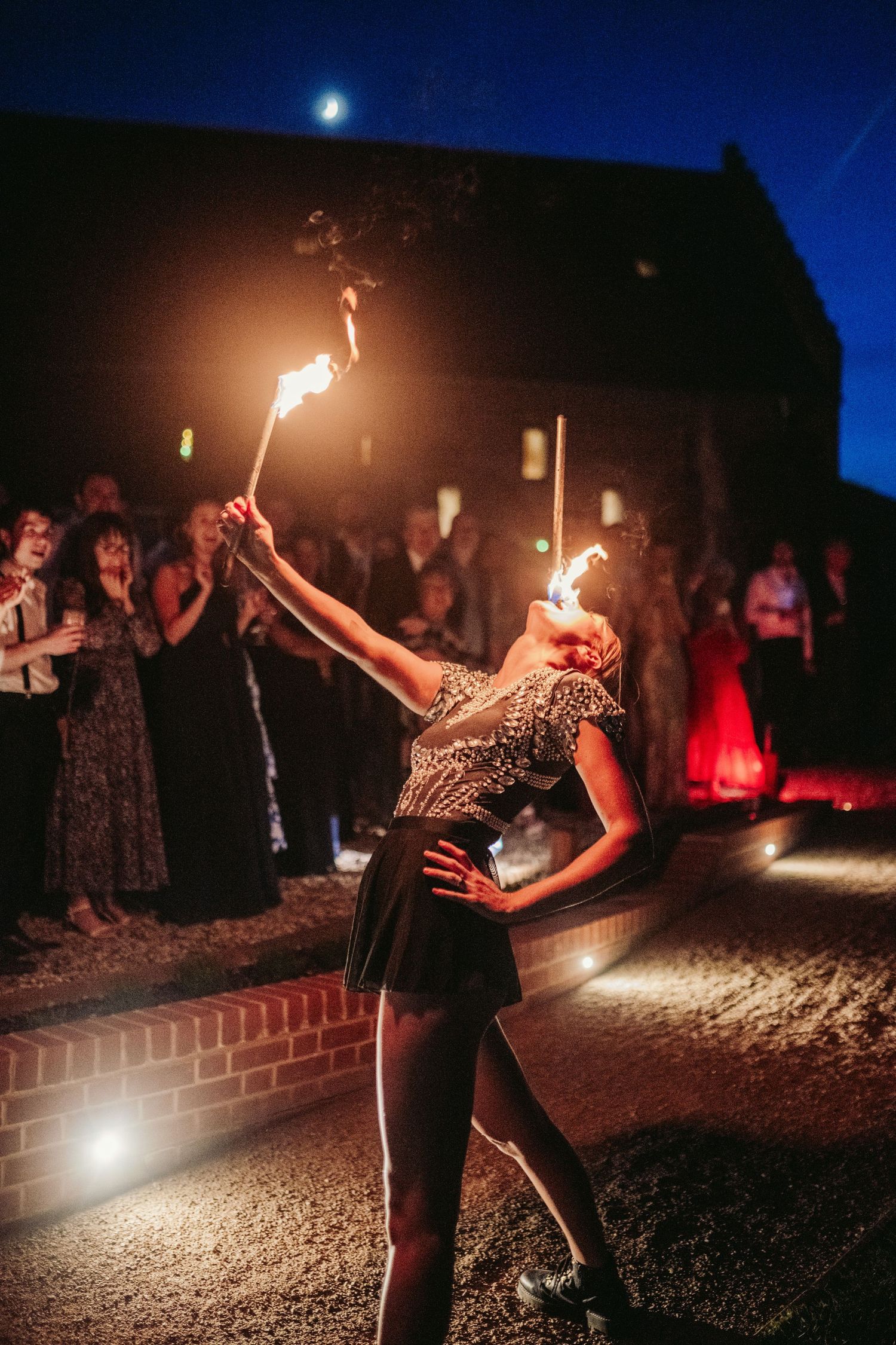 Fire dancers perform a mesmerizing nighttime display on a gravel path under a dark blue sky.
