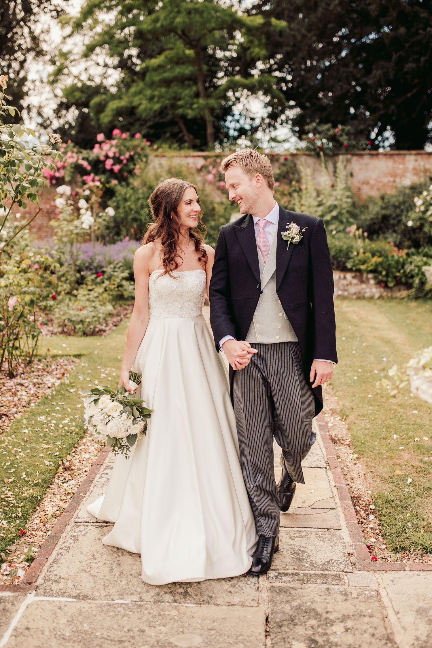 Newlyweds stroll together through a blooming garden path at their countryside wedding venue.